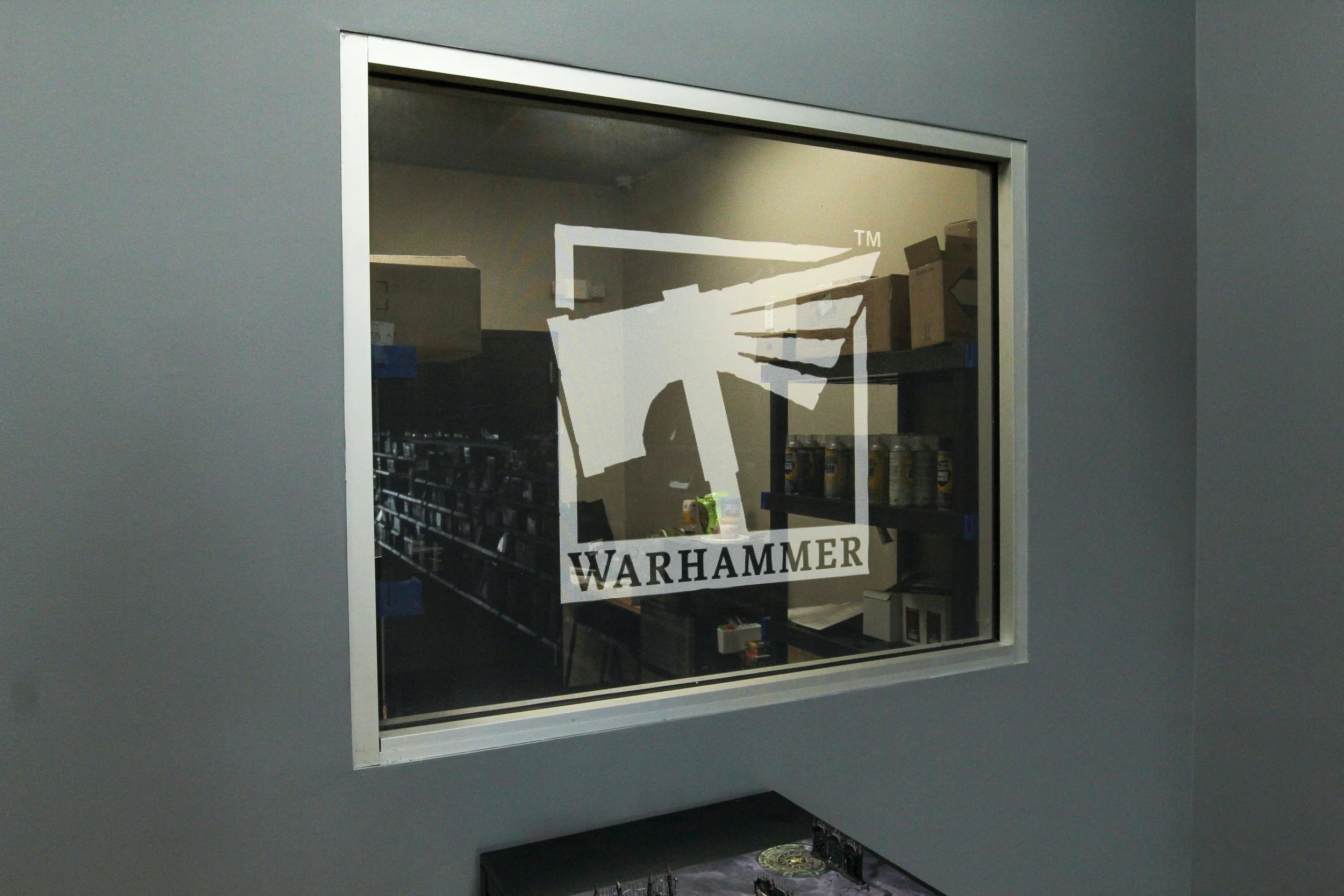A glass window with a Warhammer logo sticker on it, showing shelves with Warhammer miniatures and supplies inside. Remodeled by P&C Construction, Inc. - Chattanooga, Tn.