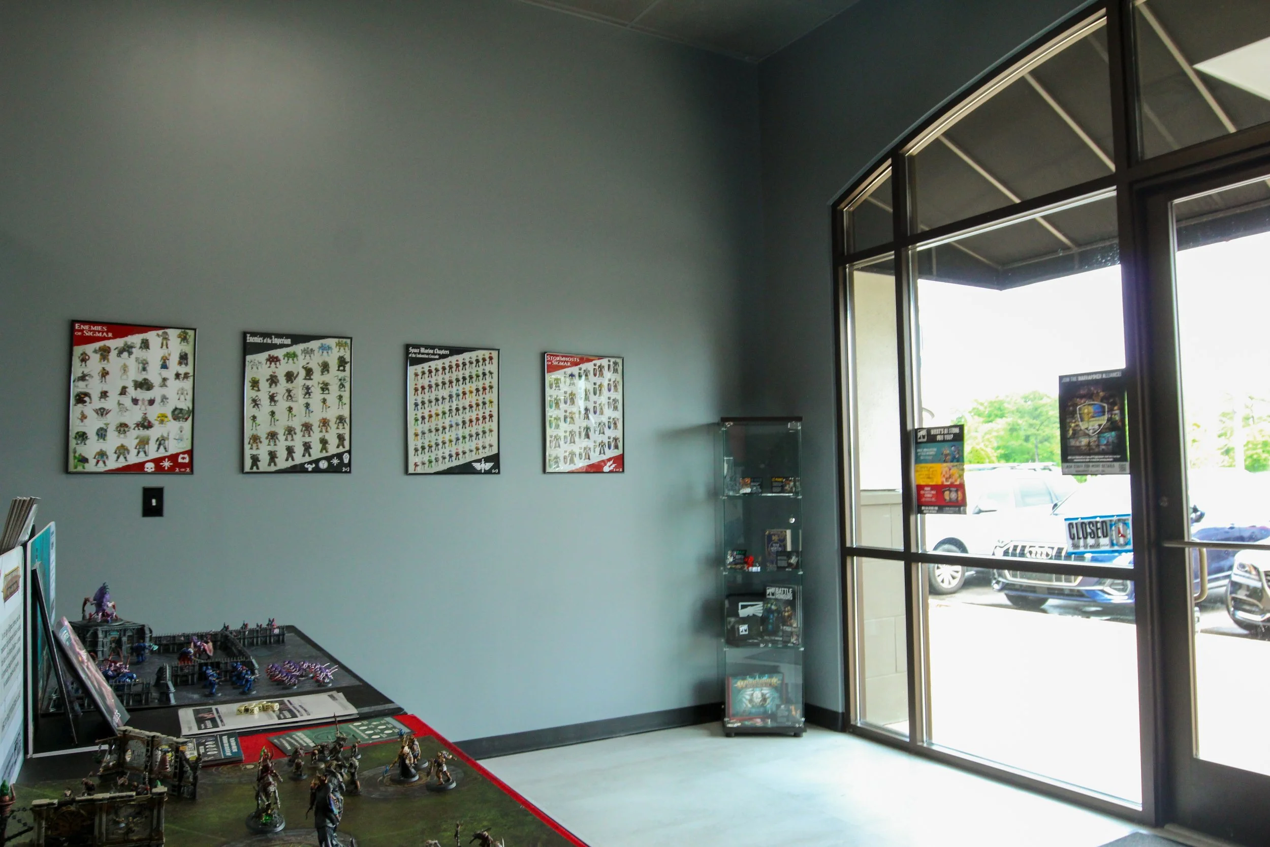 Inside a game or hobby store with miniature figurines on display, posters on the wall, and large glass windows showing parked cars outside. Remodeled by P&C Construction, Inc. - Chattanooga, Tn.
