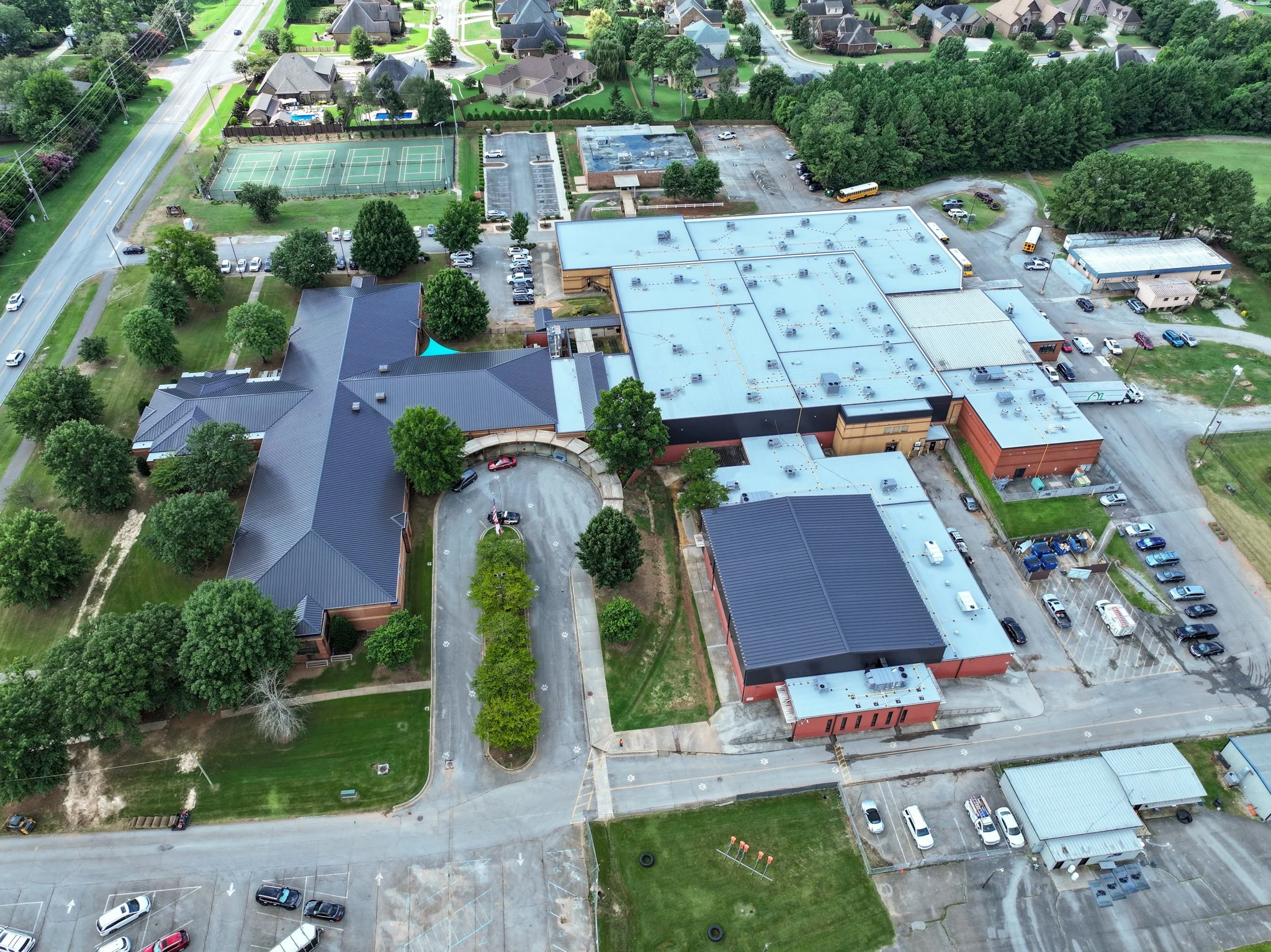 An aerial view of a suburban area featuring Discovery Middle School with a large, flat, grassy area, multiple tennis courts, a parking lot with many cars, surrounding houses, and green trees. Built by P&C Construction. 