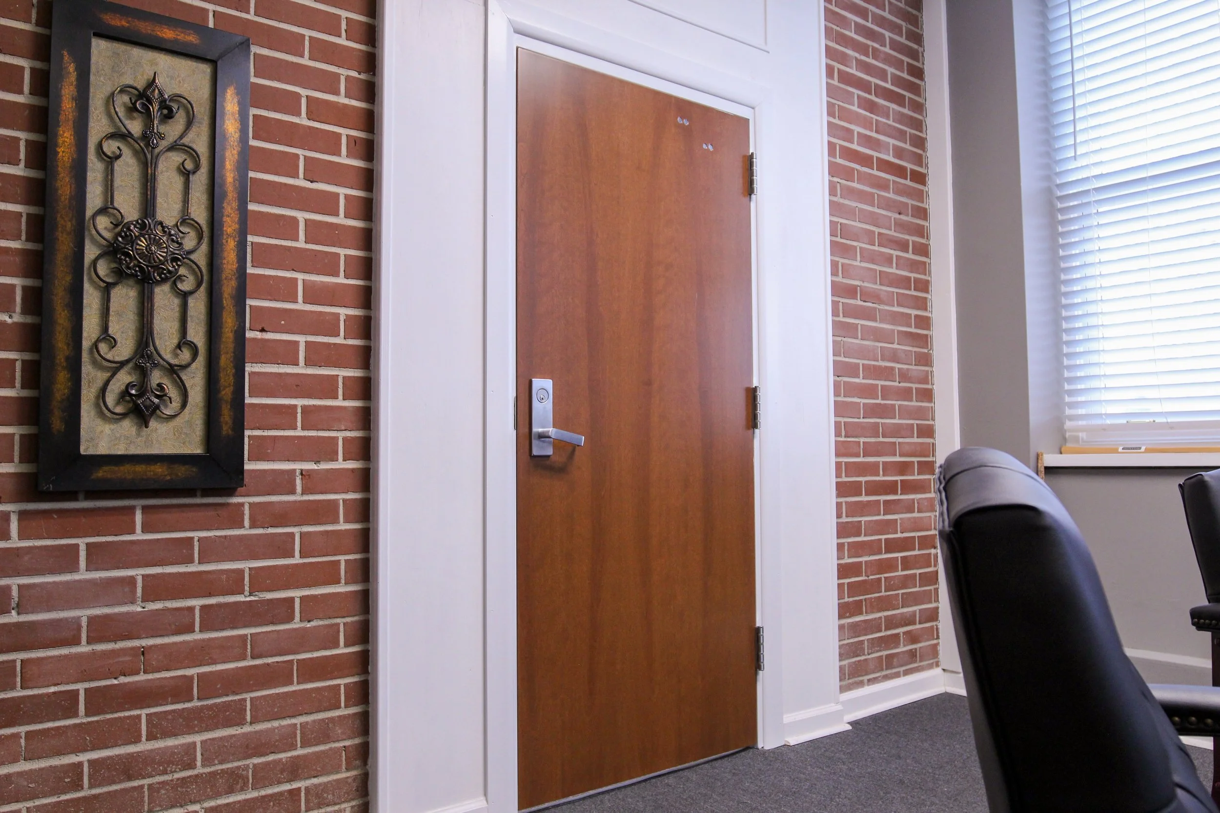 Office room with brick wall, a wooden door, black leather chairs, a window with blinds, and decorative wall art.