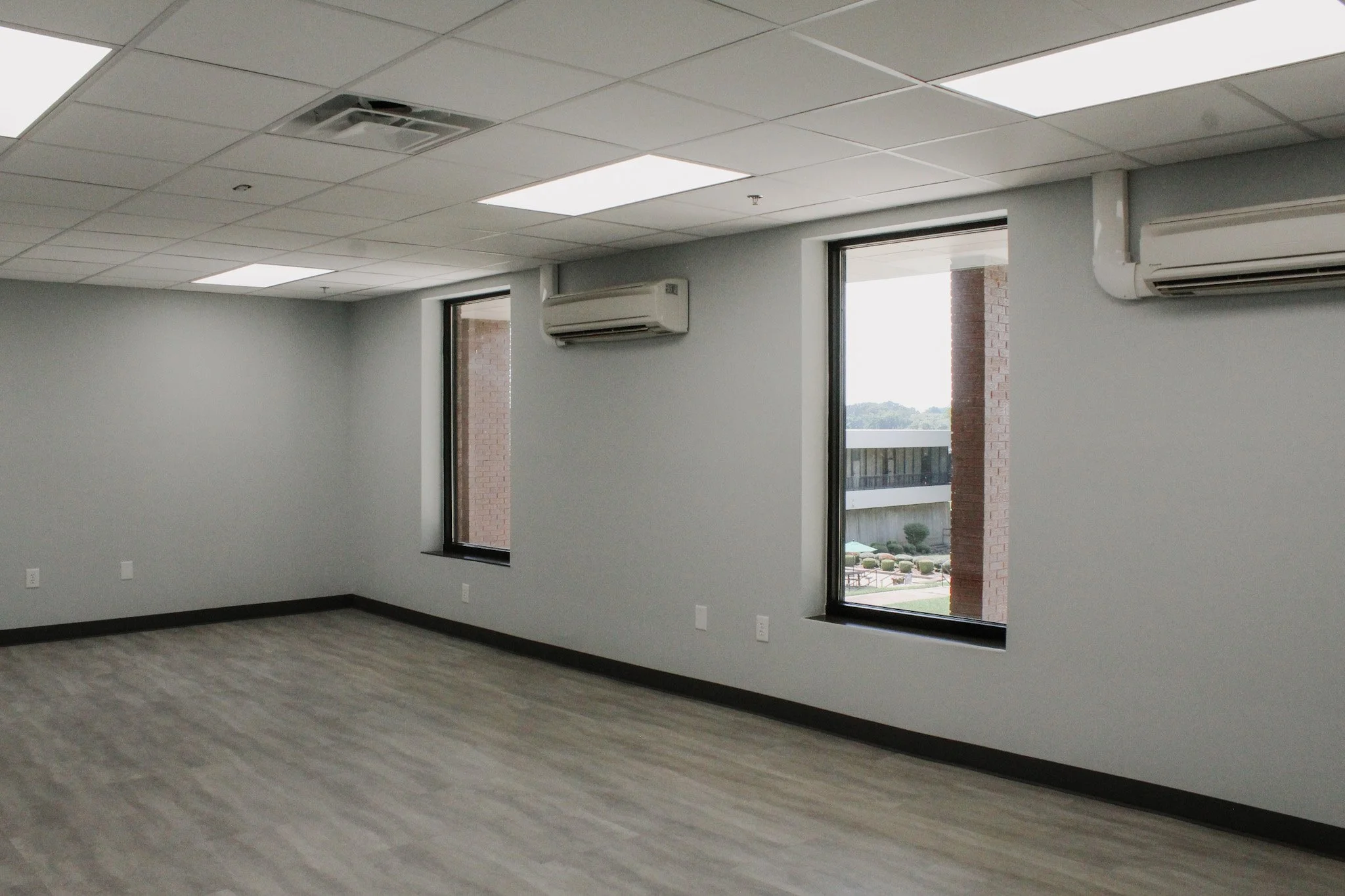 Empty office room with large windows, gray walls, ceiling lights, and air conditioning units.