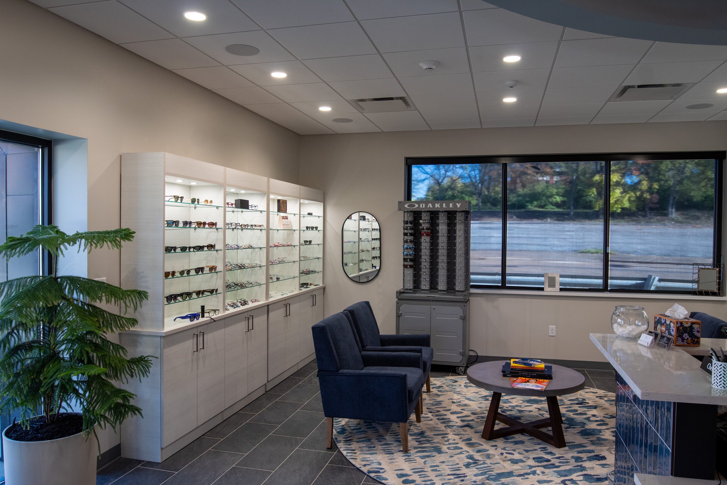 An interior of an eyewear store with display shelves full of sunglasses, a seating area with two dark blue chairs and a round table, a large window showing trees outside, and a display stand for Oakley sunglasses near the window.