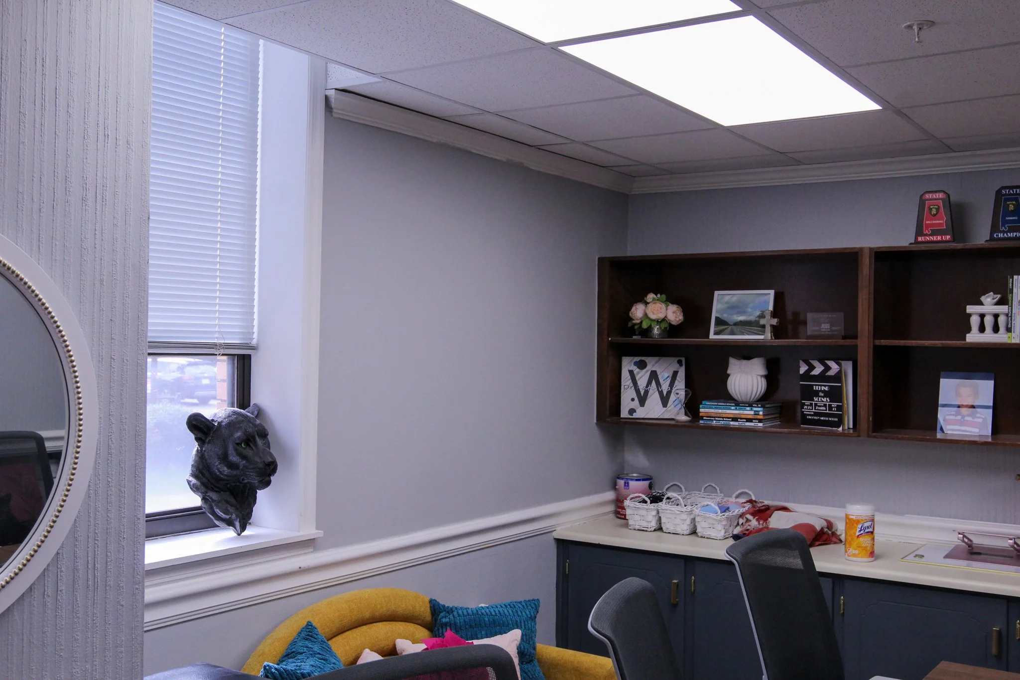 An office room with a window, a wall shelf with books and decorations, a table with miscellaneous items, and chairs. There is a yellow couch with pillows near the window and a decorative lion sculpture near the window.