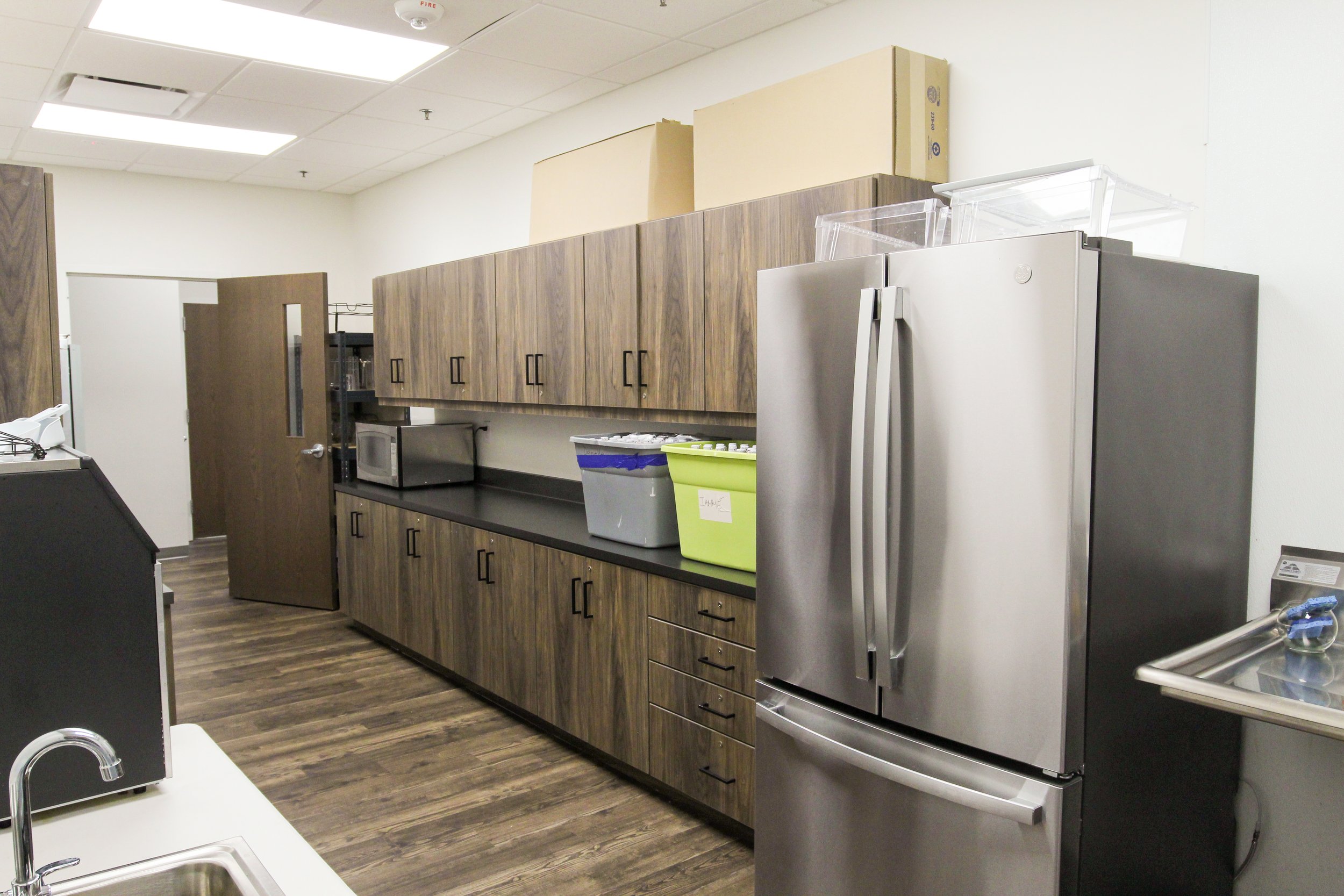 A commercial kitchen with wooden cabinets, a gray refrigerator, and a microwave on the counter. Cardboard boxes are stored on top of the cabinets. The floor is wood, and the ceiling has fluorescent lights. Design build by P&C Construction, Inc.