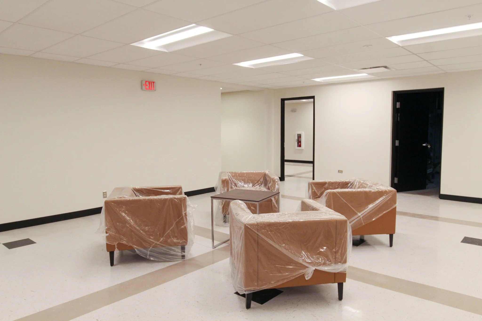 Empty waiting room with chairs covered in plastic, white walls, black baseboards, ceiling tiles, and fluorescent lights. There is a doorframe to the right, an exit sign, and a fire extinguisher mounted inside a glass case.