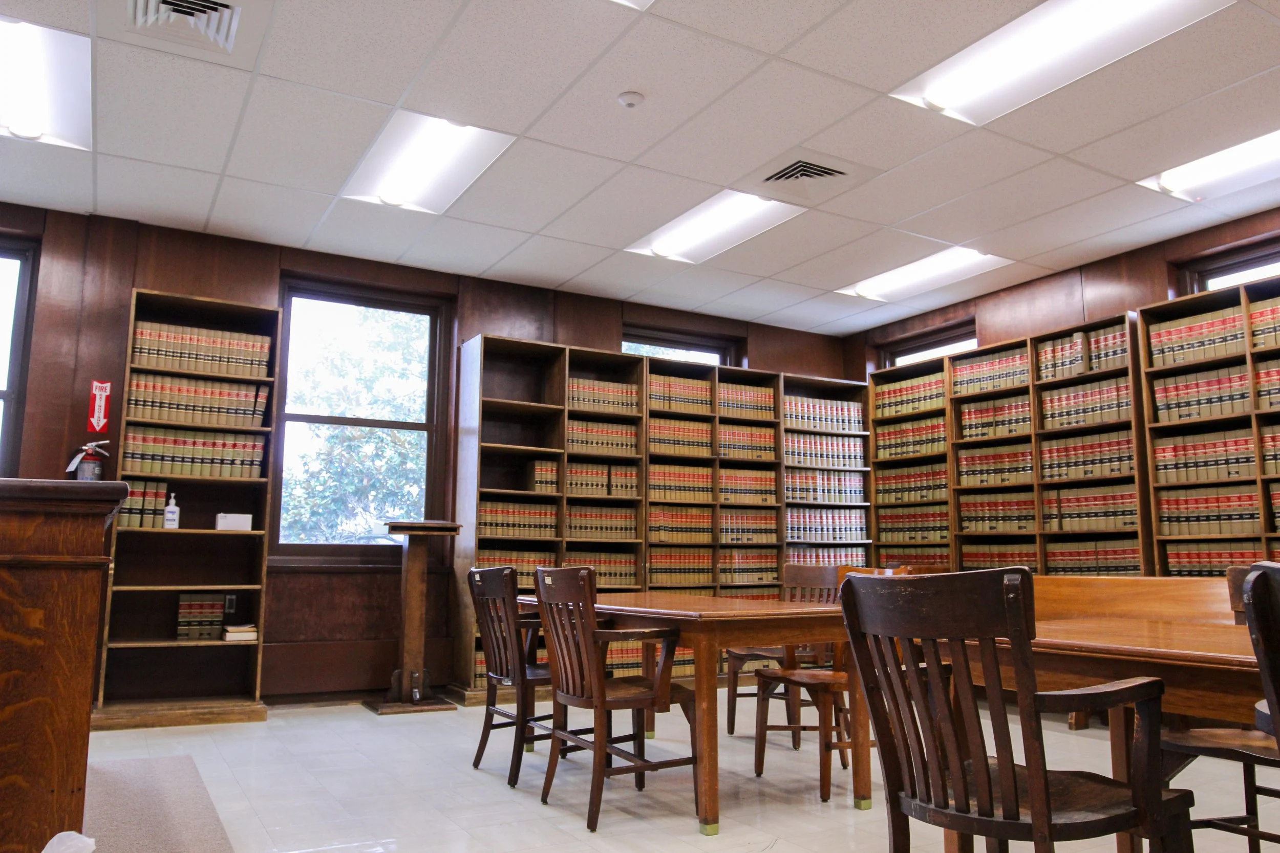 A law library with wooden shelves filled with law books, a wooden table, and chairs, two windows in the background, and fluorescent ceiling lights.