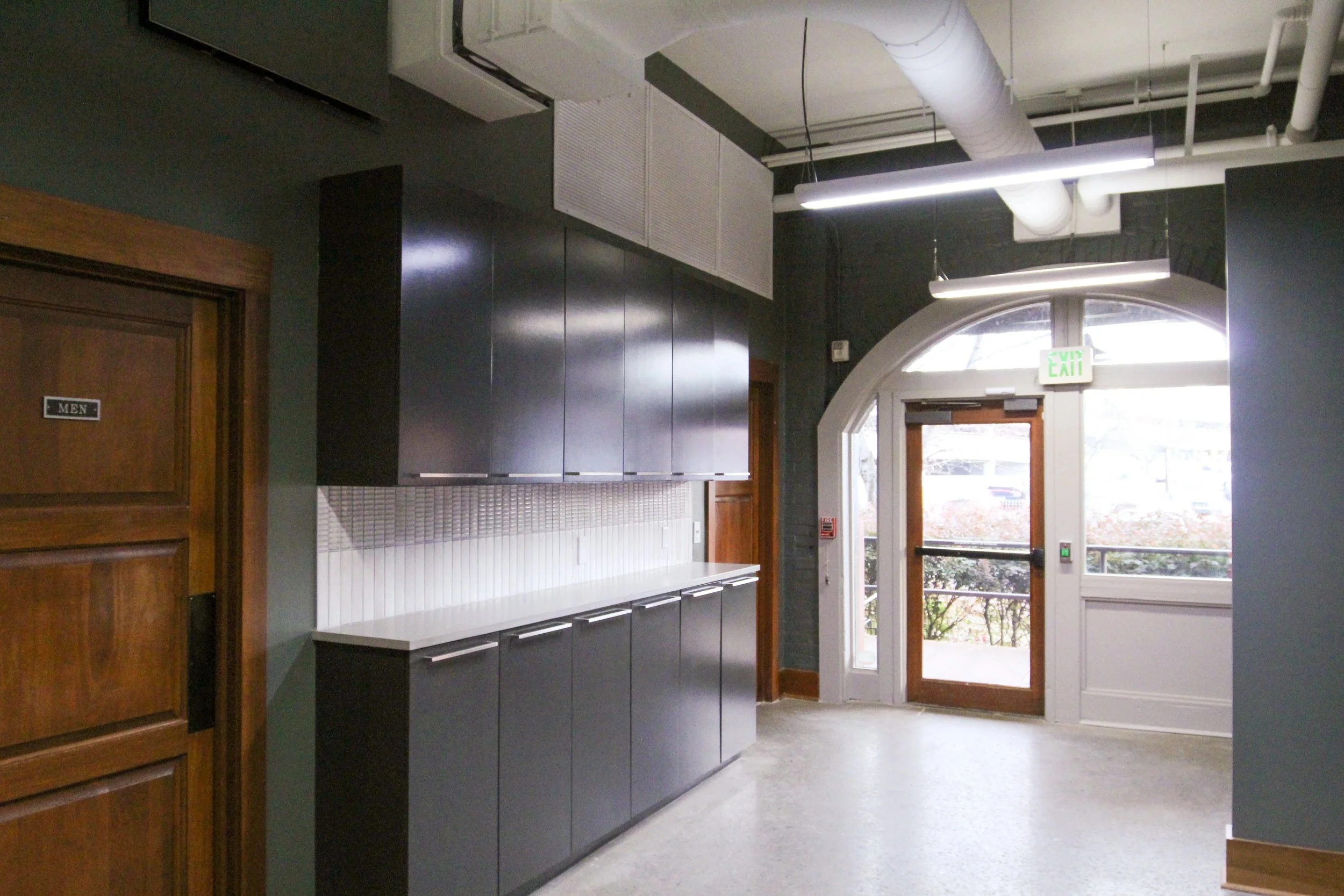 Interior of a hallway with dark cabinets, white tile backsplash, wooden doors, and glass door leading outside, with an exit sign above.