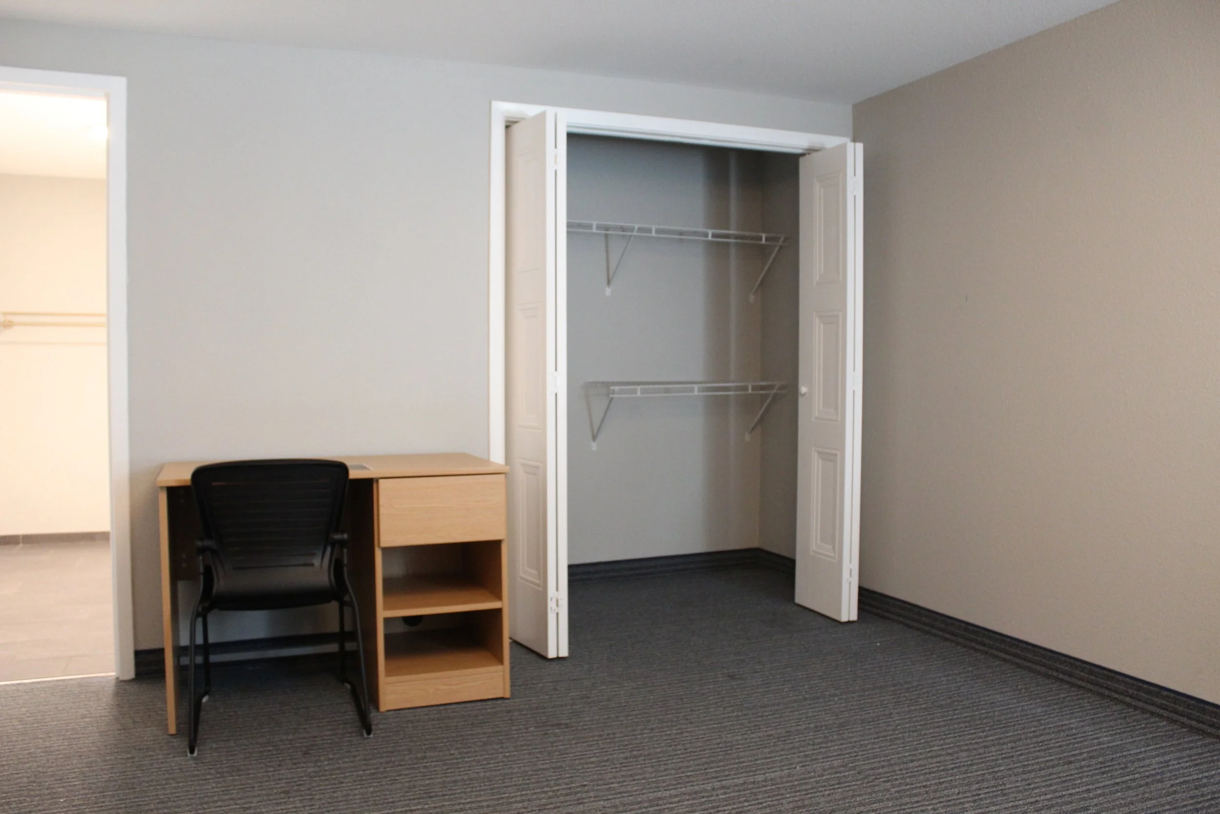 An empty bedroom corner with a closet, a small wooden desk, and an office chair. The closet has double doors, one open, with wire shelves inside. The room has gray walls, a gray carpet, and a white ceiling.
