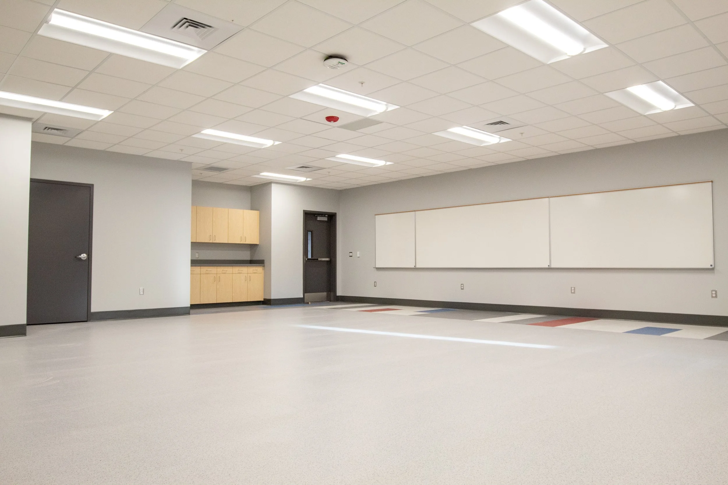Empty classroom with whiteboard, cabinets, door, and ceiling lights.