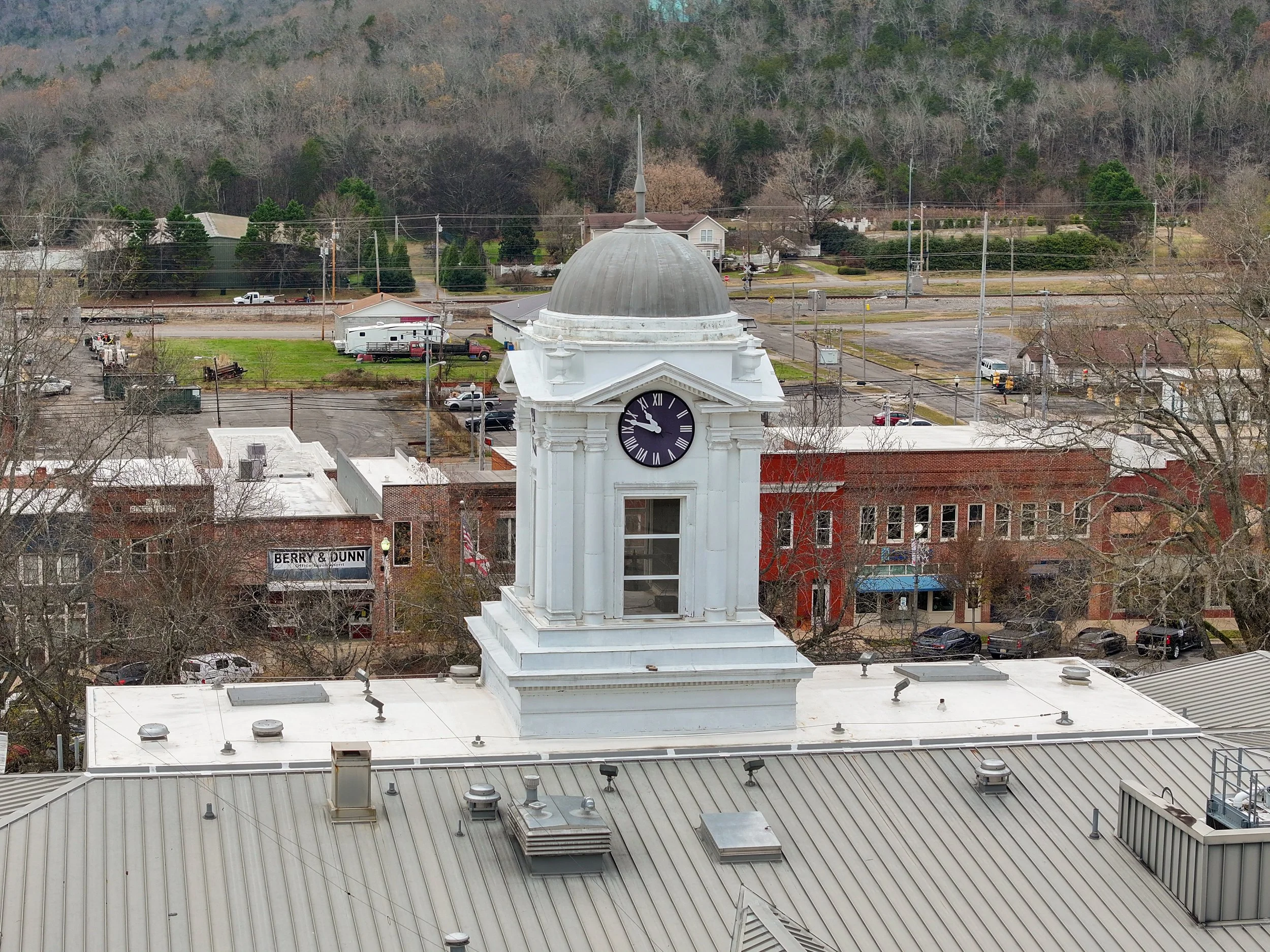 A clock tower with a black face and white Roman numerals on a white building, situated on a rooftop in a small town with commercial buildings, parked cars, trees, and a landscape with hills in the background.