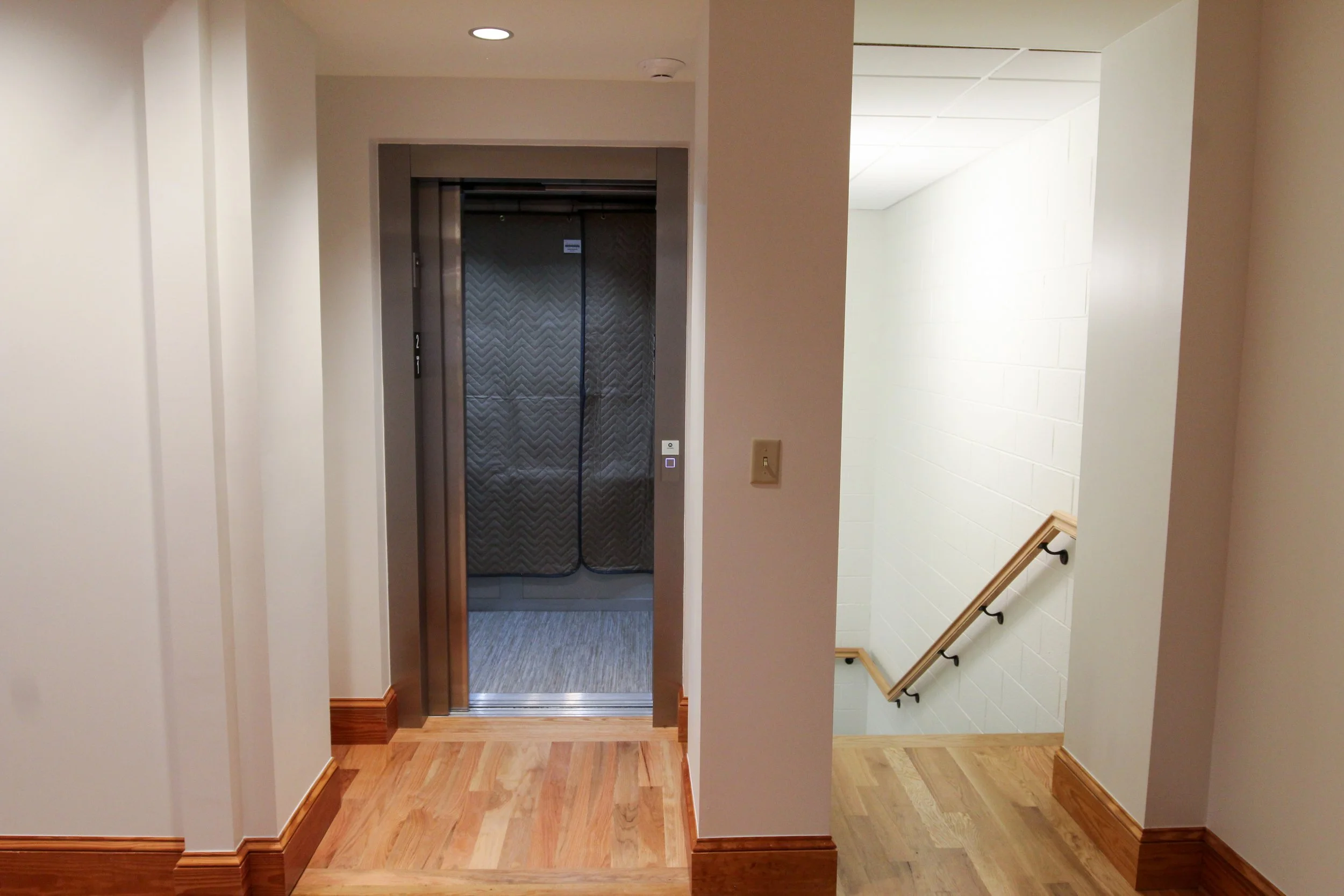 An indoor scene with an elevator on the left and a stairway with a handrail on the right, in a building corridor with light-colored walls and wooden trim.