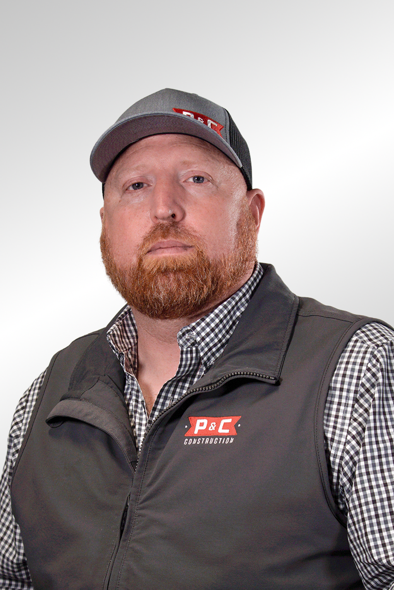 A man with a red beard wearing a gray and black cap and a black vest over a checkered shirt, standing against a plain light gray background.