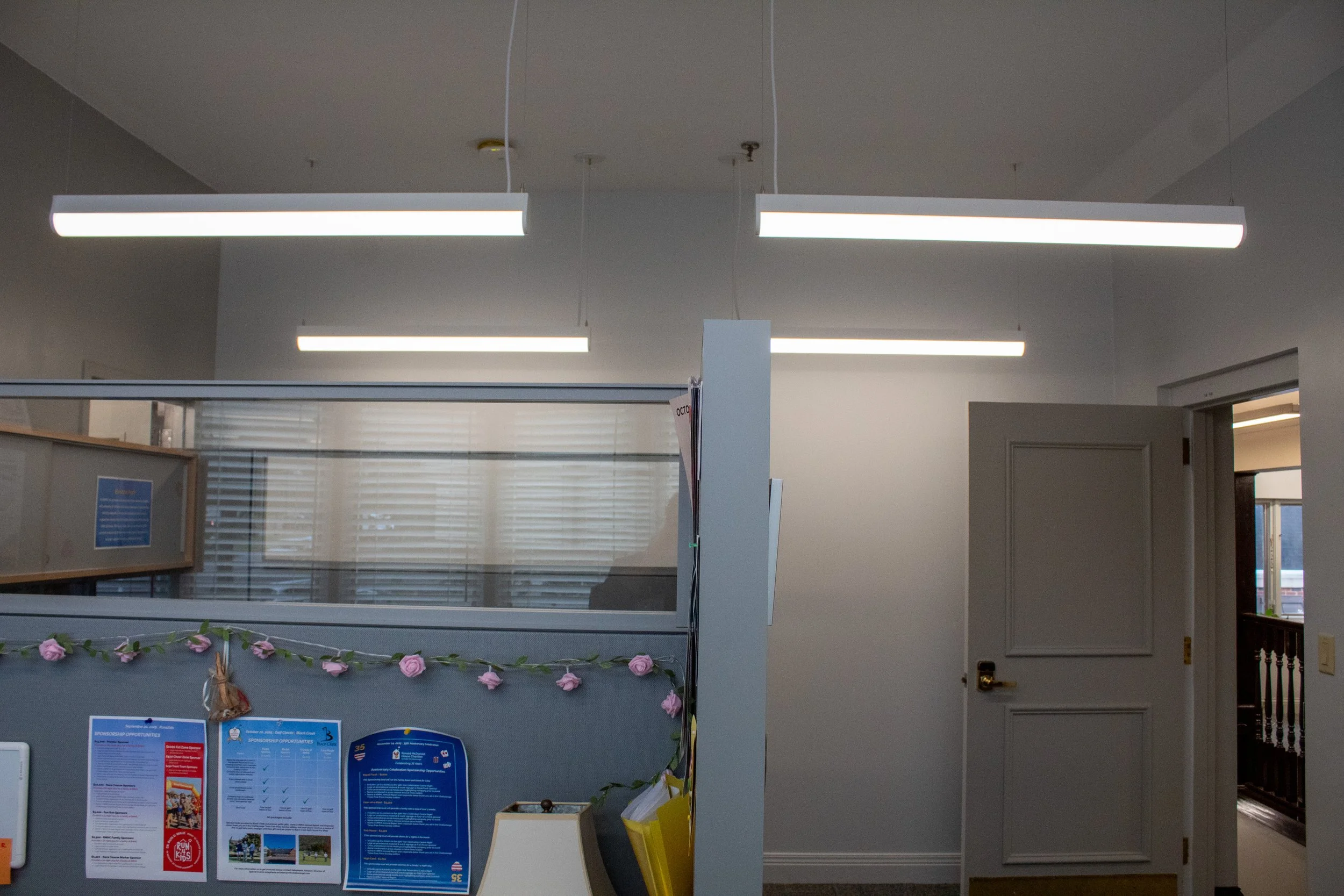 Office cubicle with decorated garland of pink flowers, posters, and informational materials, illuminated by overhead fluorescent lights, with a partially open door leading to a hallway.