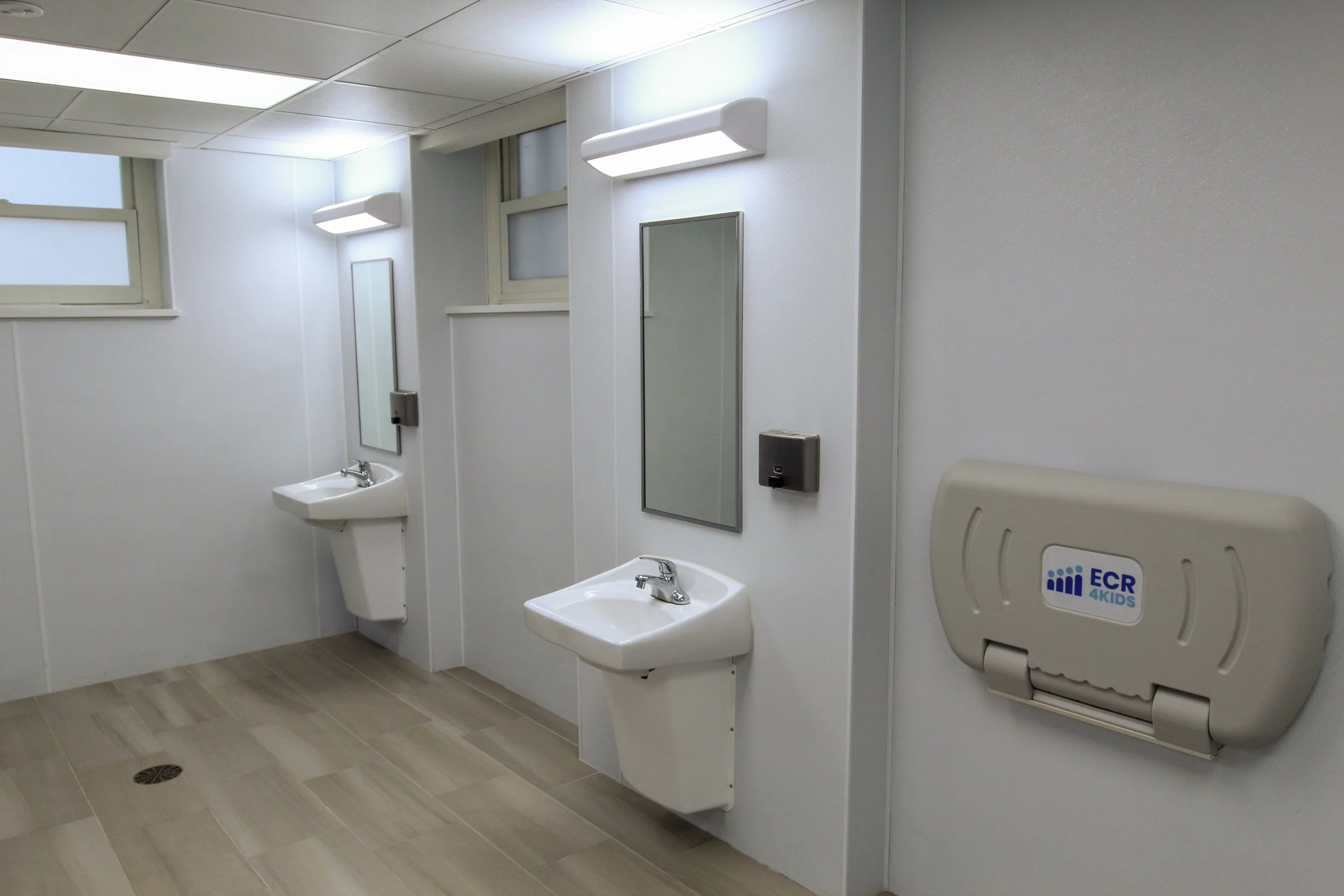 Public men's restroom with two sinks, mirrored medicine cabinets, wall-mounted hand dryers, a paper towel dispenser, and small windows near the ceiling.