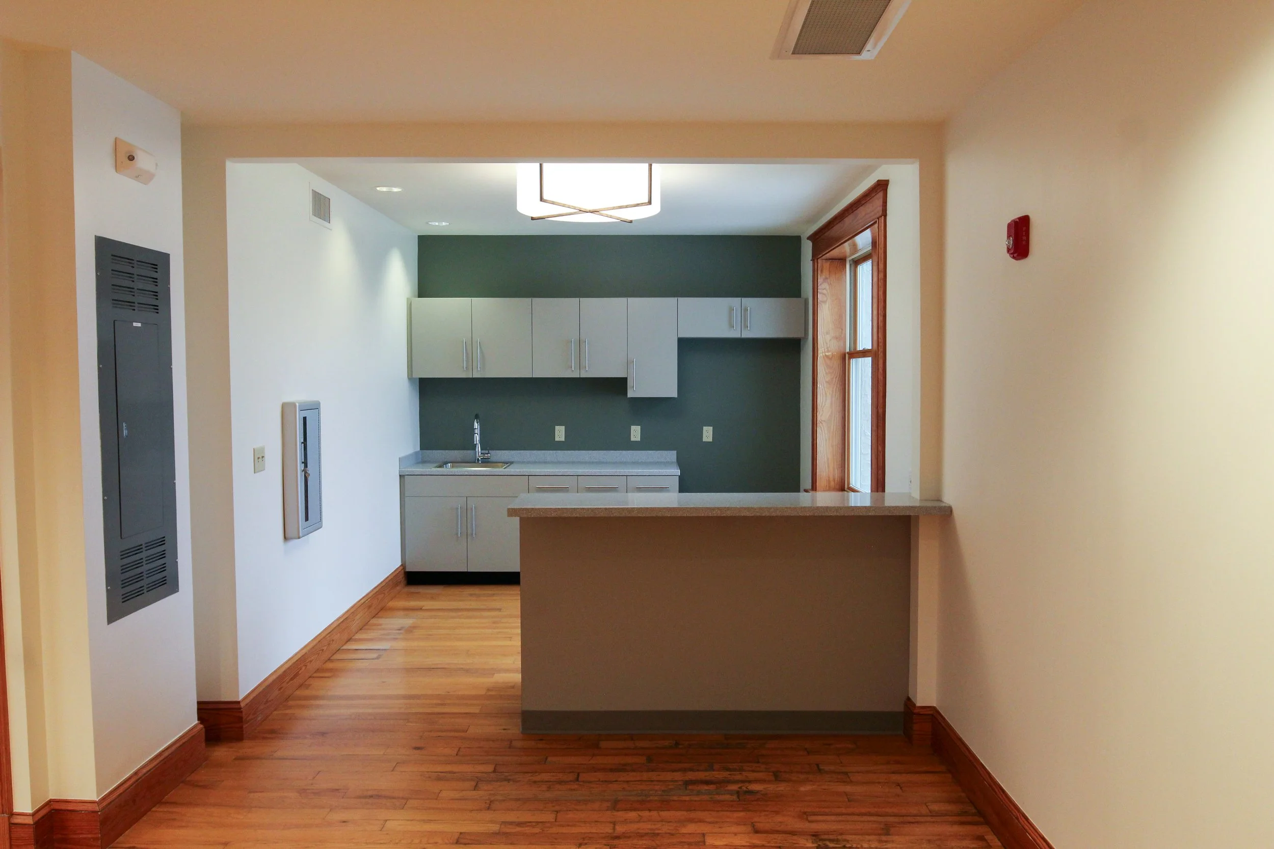 An empty kitchen with grey cabinets, a sink, wooden trim and flooring, white walls, and a green accent wall. There is a window with a wooden frame and a ceiling light fixture.