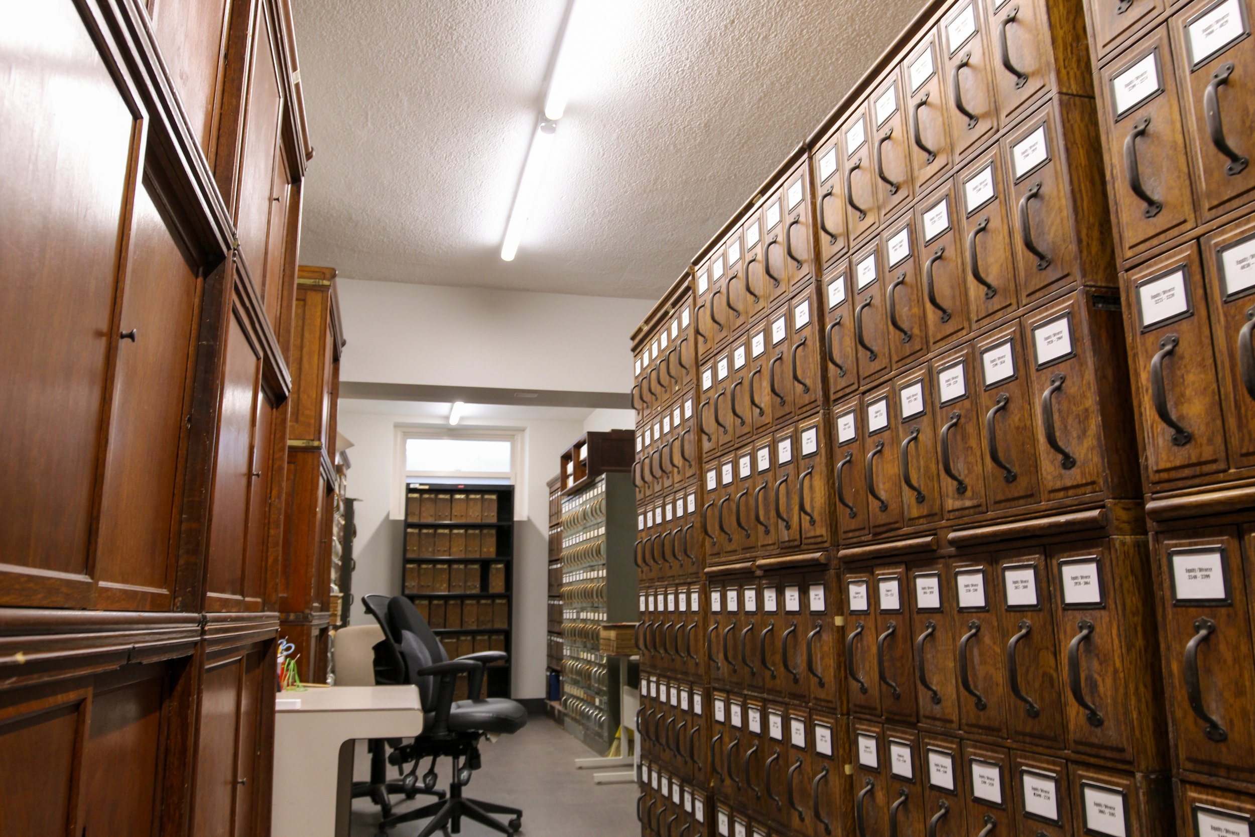 Room filled with wooden filing cabinets and shelves containing binders and folders, with chairs and a desk in the corner.