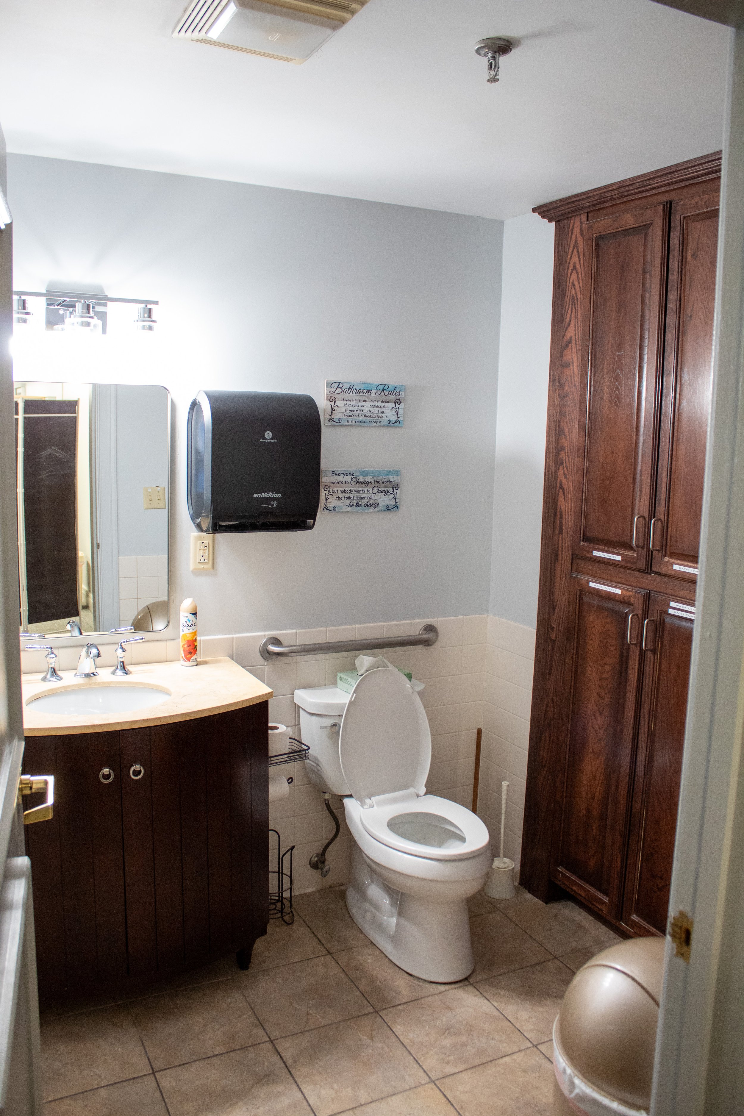 Bathroom with a toilet, a wooden cabinet, a sink with a mirror, a wall-mounted paper towel dispenser, and decorative signs on the wall.