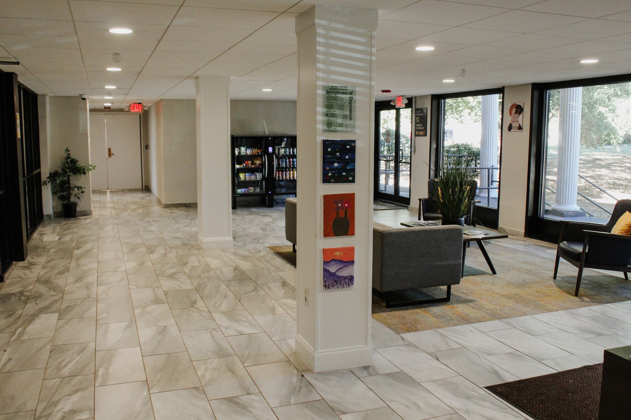 Interior of a lobby or waiting area with marble flooring, large windows, chairs, a plant, a vending machine, and artwork on the wall.