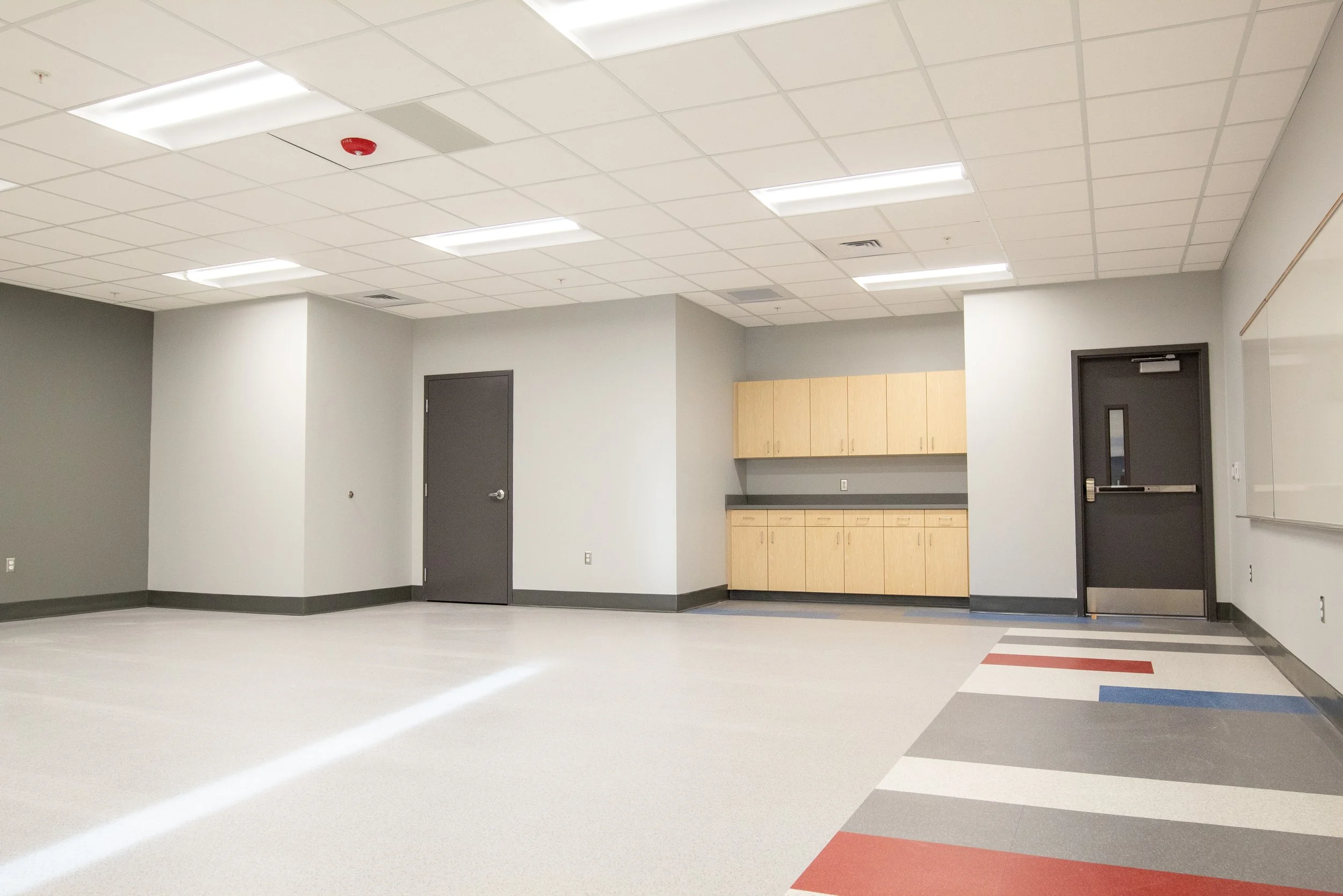 Empty classroom or meeting room with gray doors, a sink, and cabinets, and a colorful patterned floor.