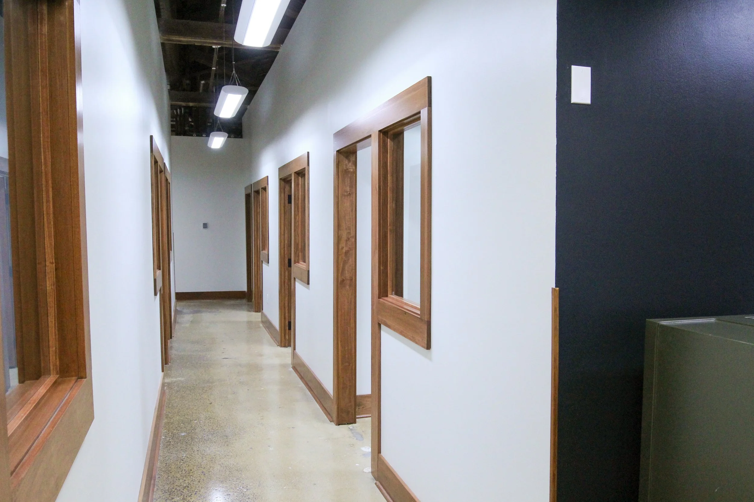 A hallway with white walls, wooden doorframes, and a polished concrete floor. Several doors line the hallway, and modern light fixtures hang from the ceiling.