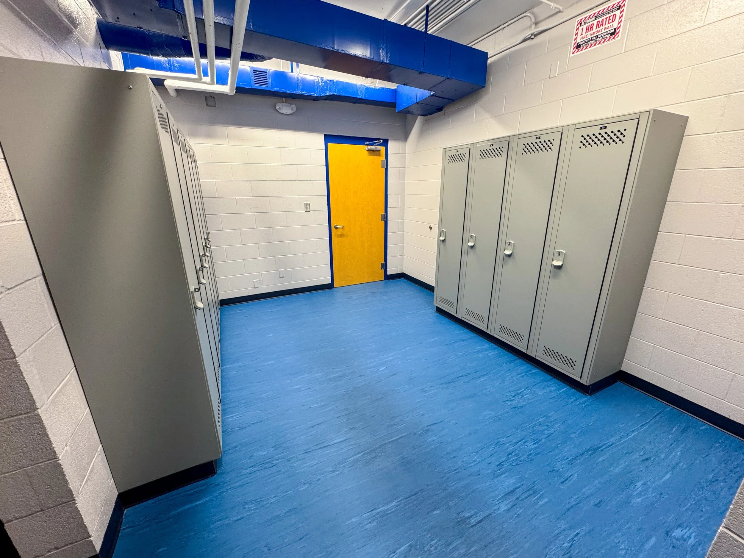 A room with blue floor and white brick walls, featuring gray lockers and a yellow door with a blue frame. Above, there is a blue duct and a red and white fire safety sign.
