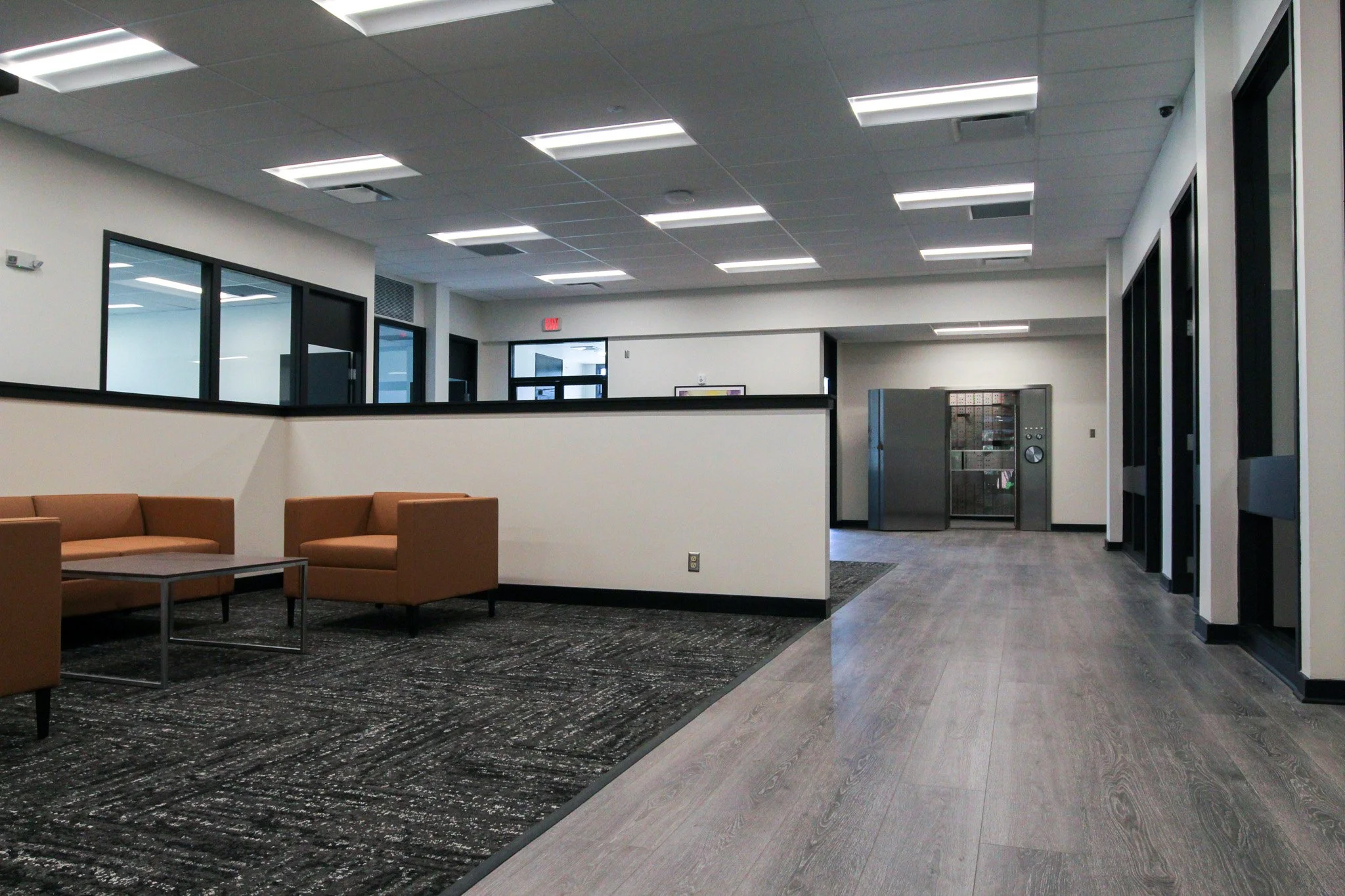 An indoor seating area with brown cushioned chairs and a small coffee table, modern gray carpeting, and a section of grayish wooden flooring. The background features large glass windows and doors, with a hallway and elevator visible further back in a