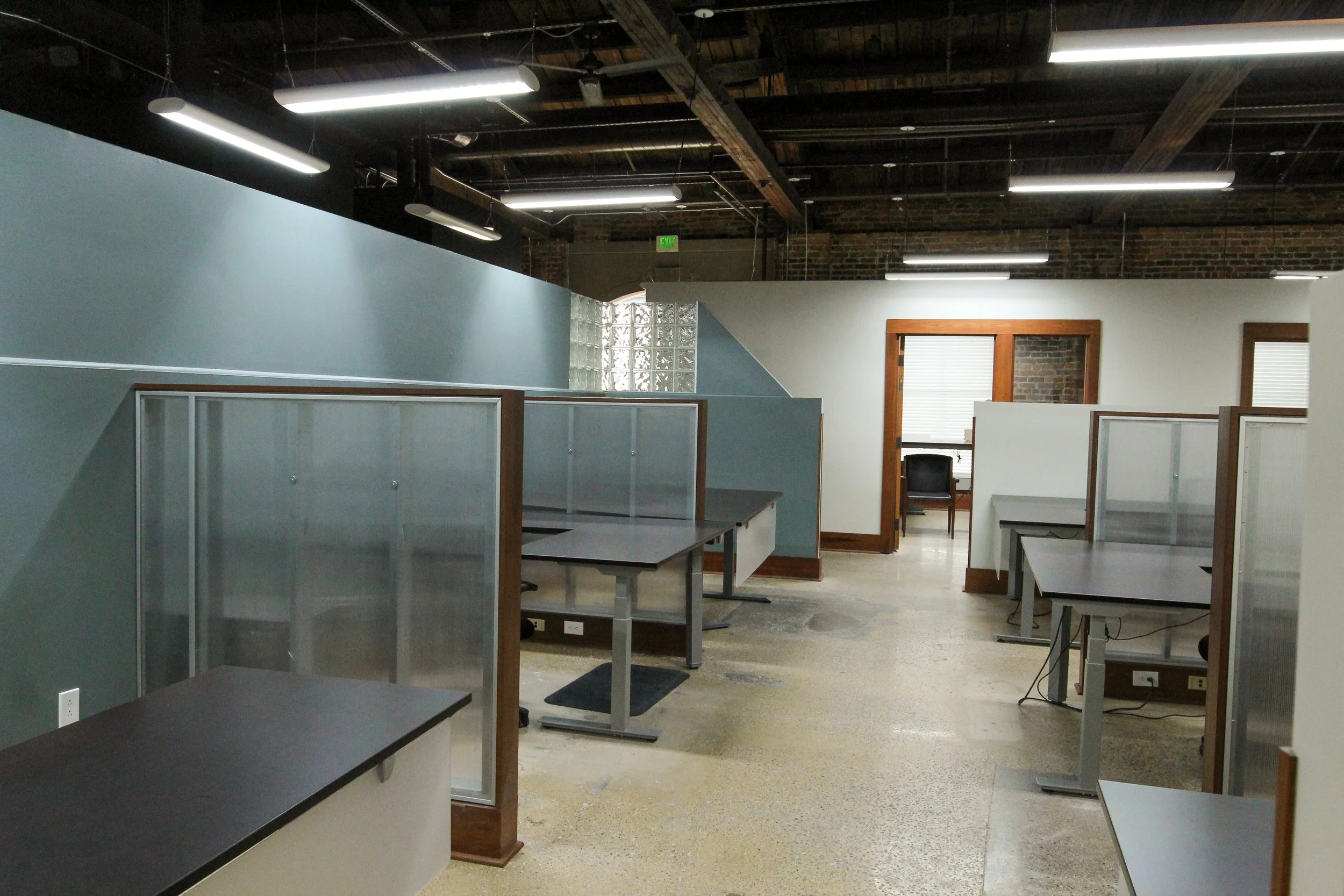 Empty office space with cubicles, desks, and chairs, illuminated by fluorescent lighting, with brick walls and windows.