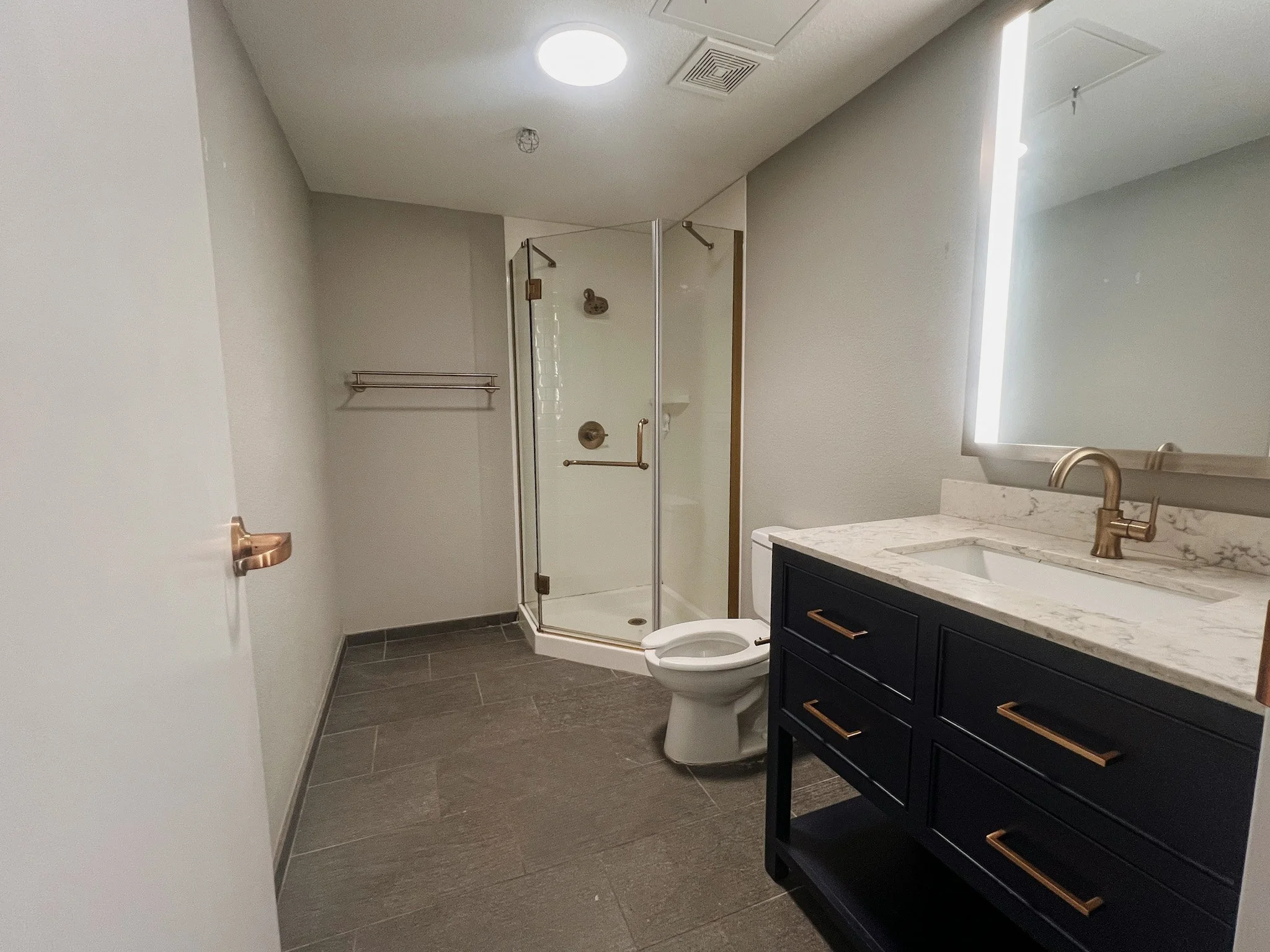 Modern bathroom with a glass shower stall, white toilet, dark blue vanity with a marble countertop, and a large illuminated mirror. Remodeled by P&C Construction, Inc. - Chattanooga, Tn.
