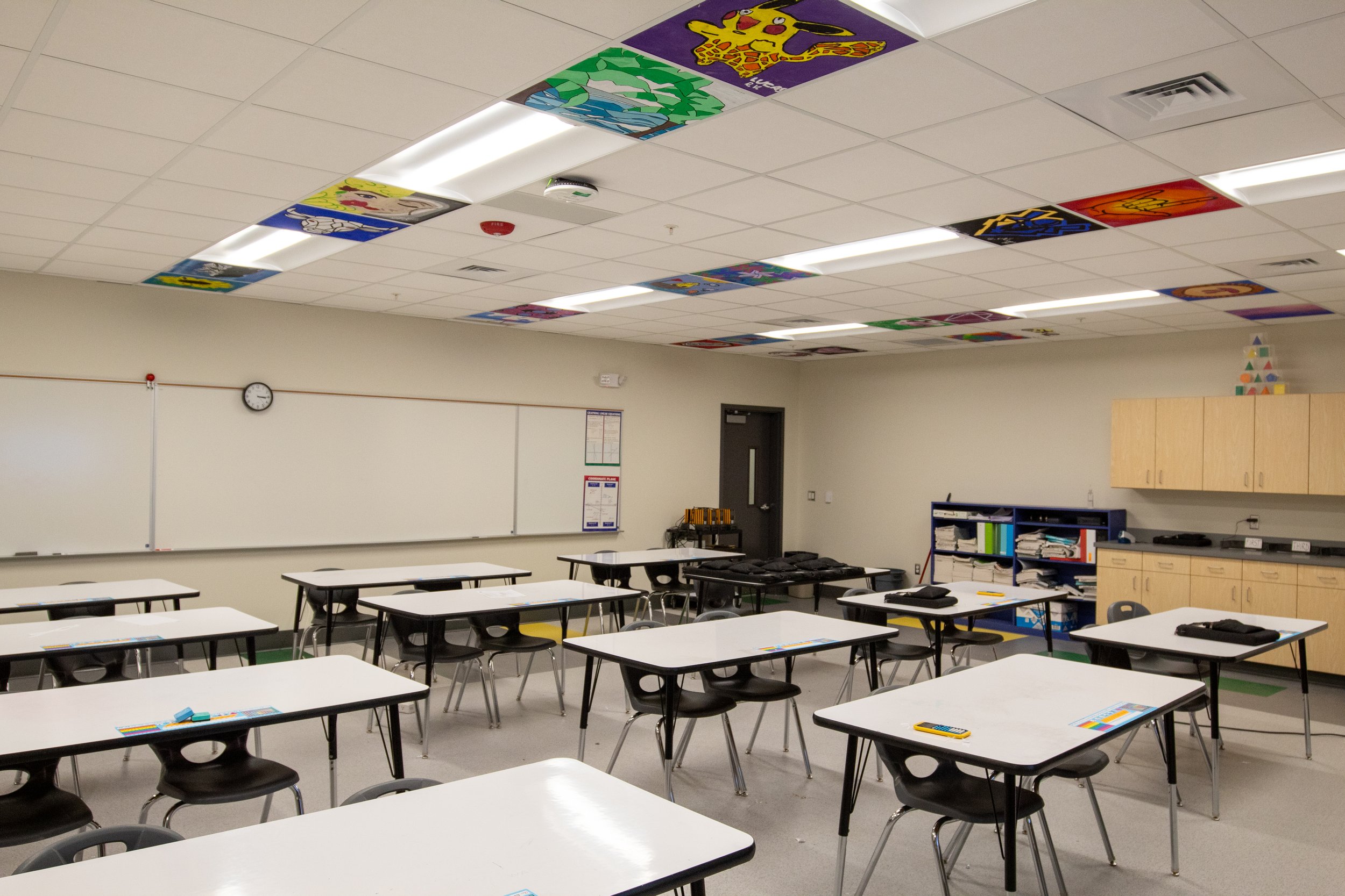 Empty classroom with white desks, black chairs, a whiteboard, and colorful artwork of cartoon characters on the ceiling.