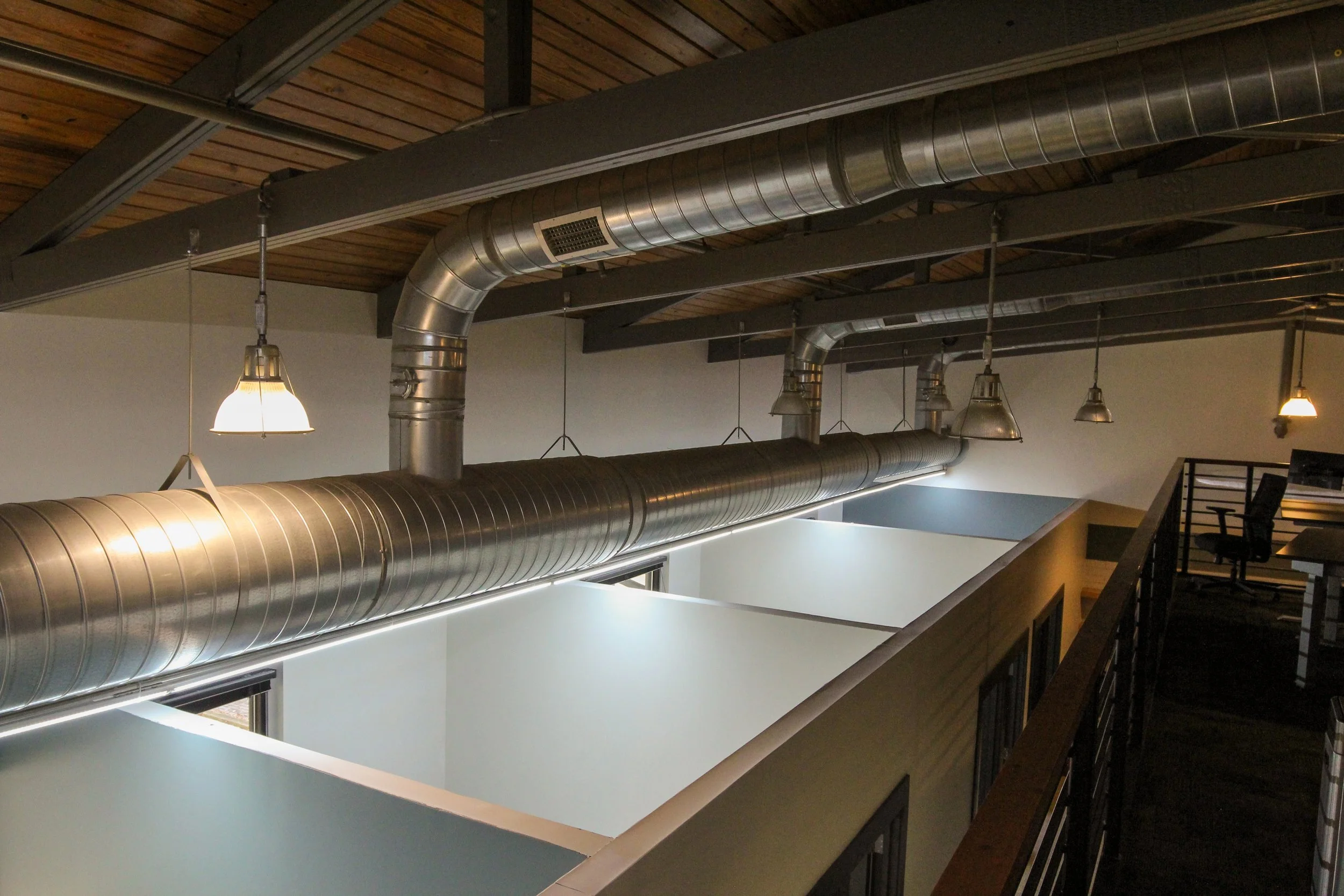 Office with suspended metallic ductwork, hanging pendant lights, and a wooden ceiling, featuring a balcony with black railing.