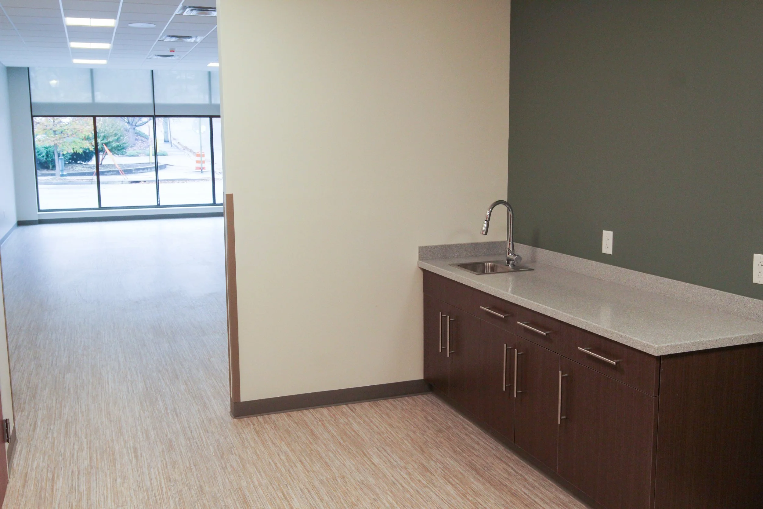 Empty office kitchenette with dark wood cabinets, a gray countertop, and a stainless steel sink, with a large windowed area in the background.