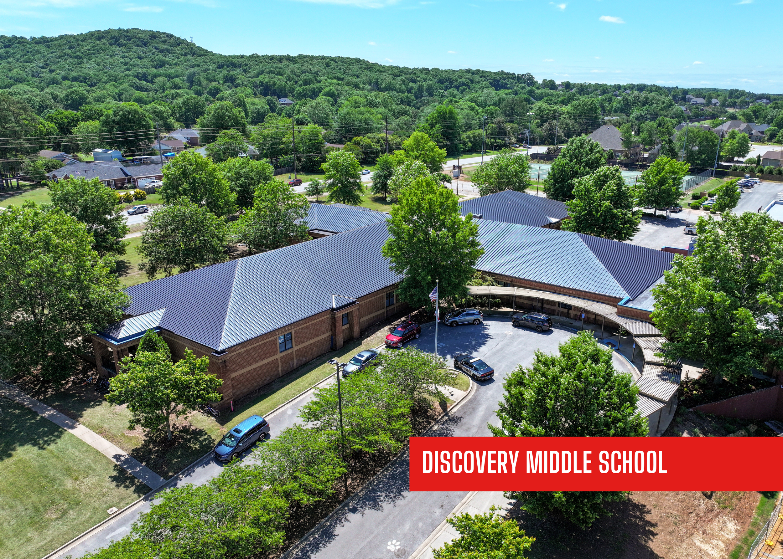 Aerial view of Discovery Middle School, a building with a dark gray metal roof, surrounded by trees, parking lots, and residential neighborhoods with a hilly, forested background. Roof remodeled by P&C Construction, Inc. - Chattanooga, Tn.