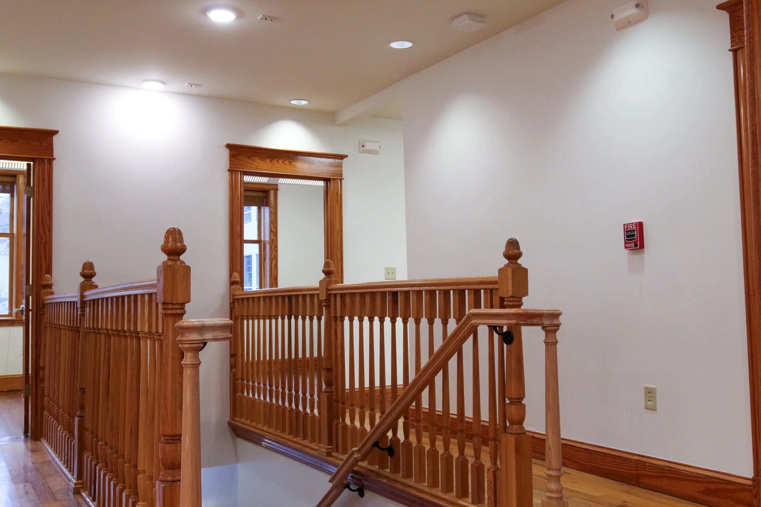 Interior of a building with white walls, wooden railing, and two windows. Ceiling has recessed lighting and smoke detectors.