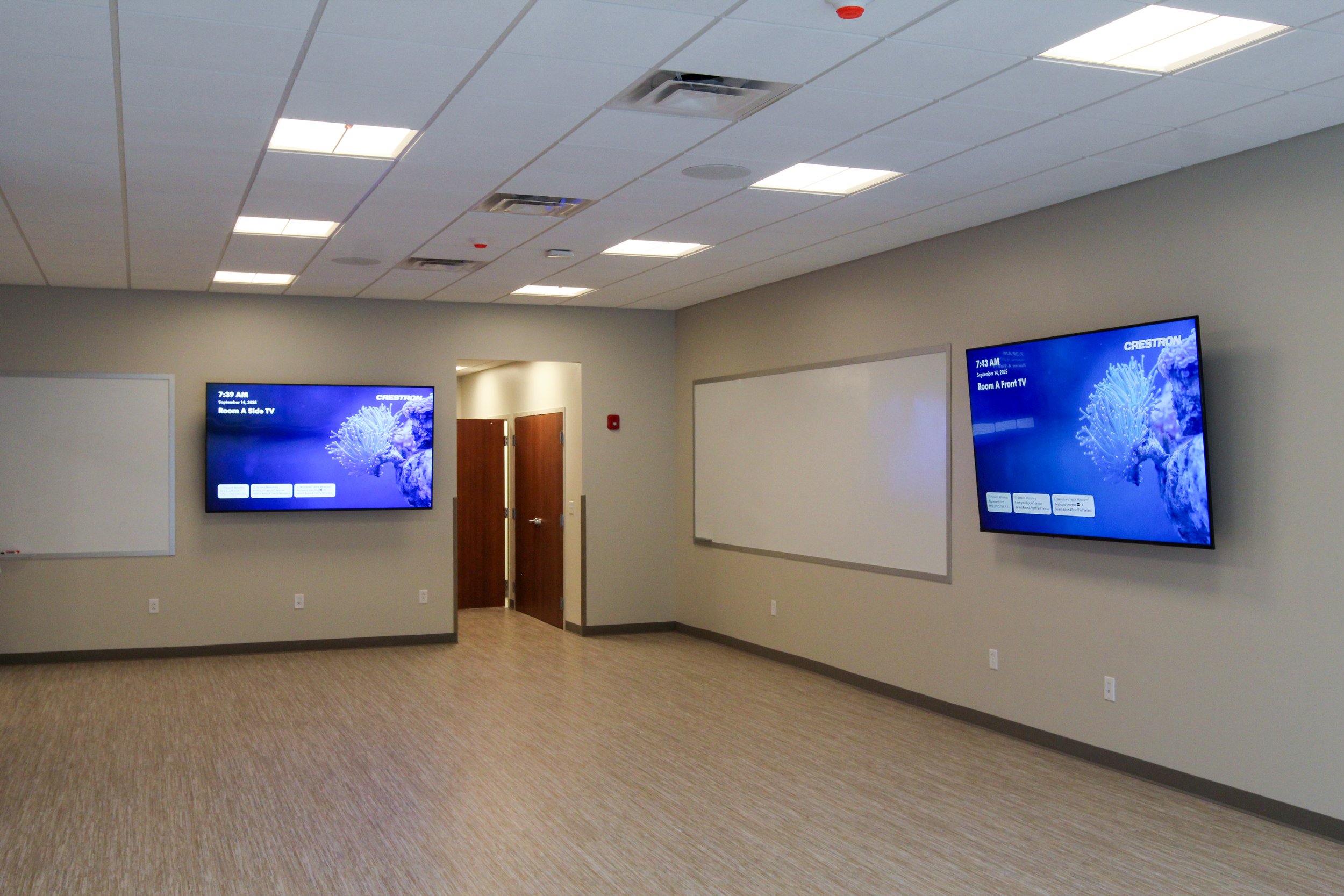 Empty classroom with two wall-mounted screens displaying blue aquatic images, two whiteboards, wooden doors, and ceiling lights.