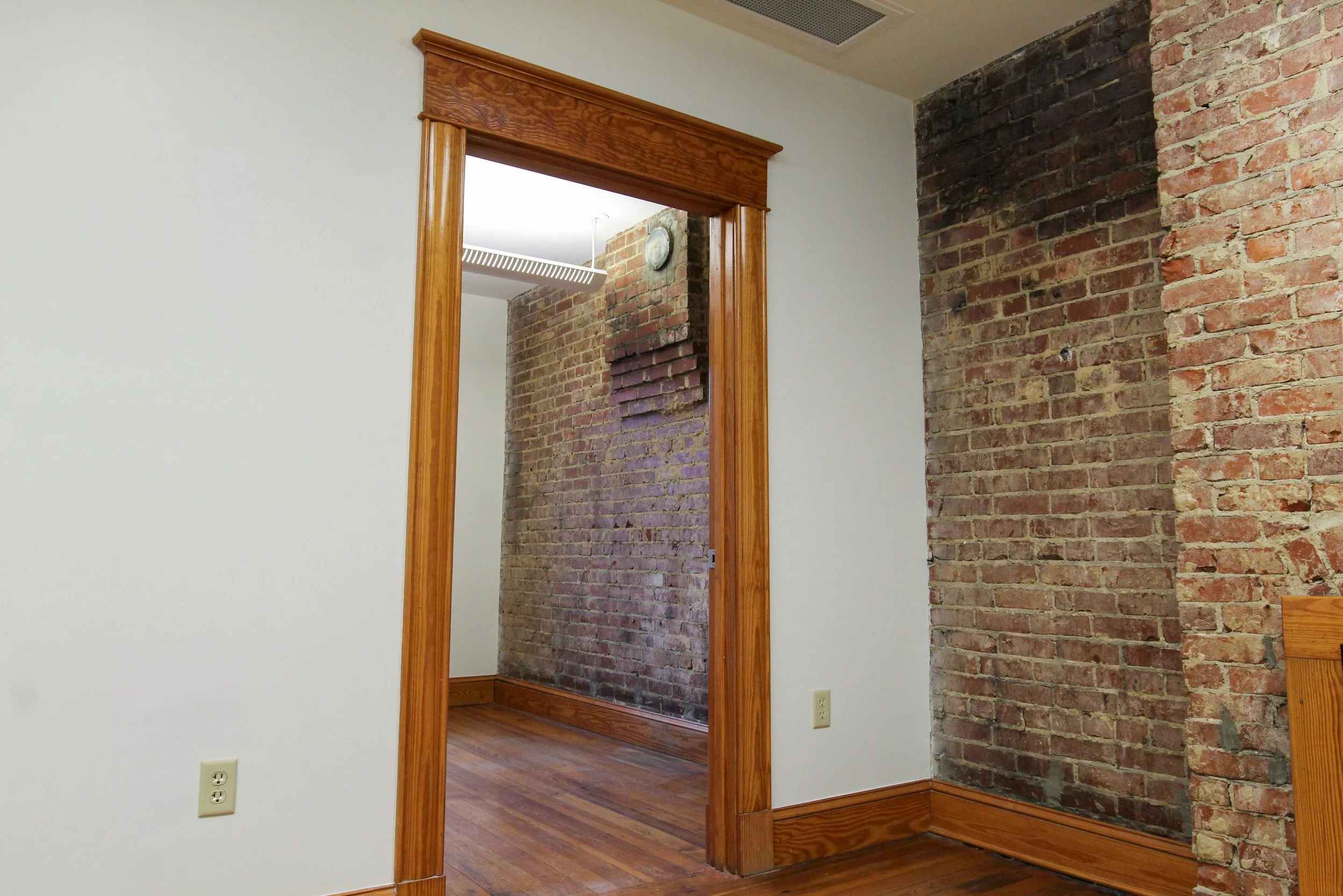 Interior of a room with brick walls, wooden trim around the doorway, wood flooring, and a ceiling vent.
