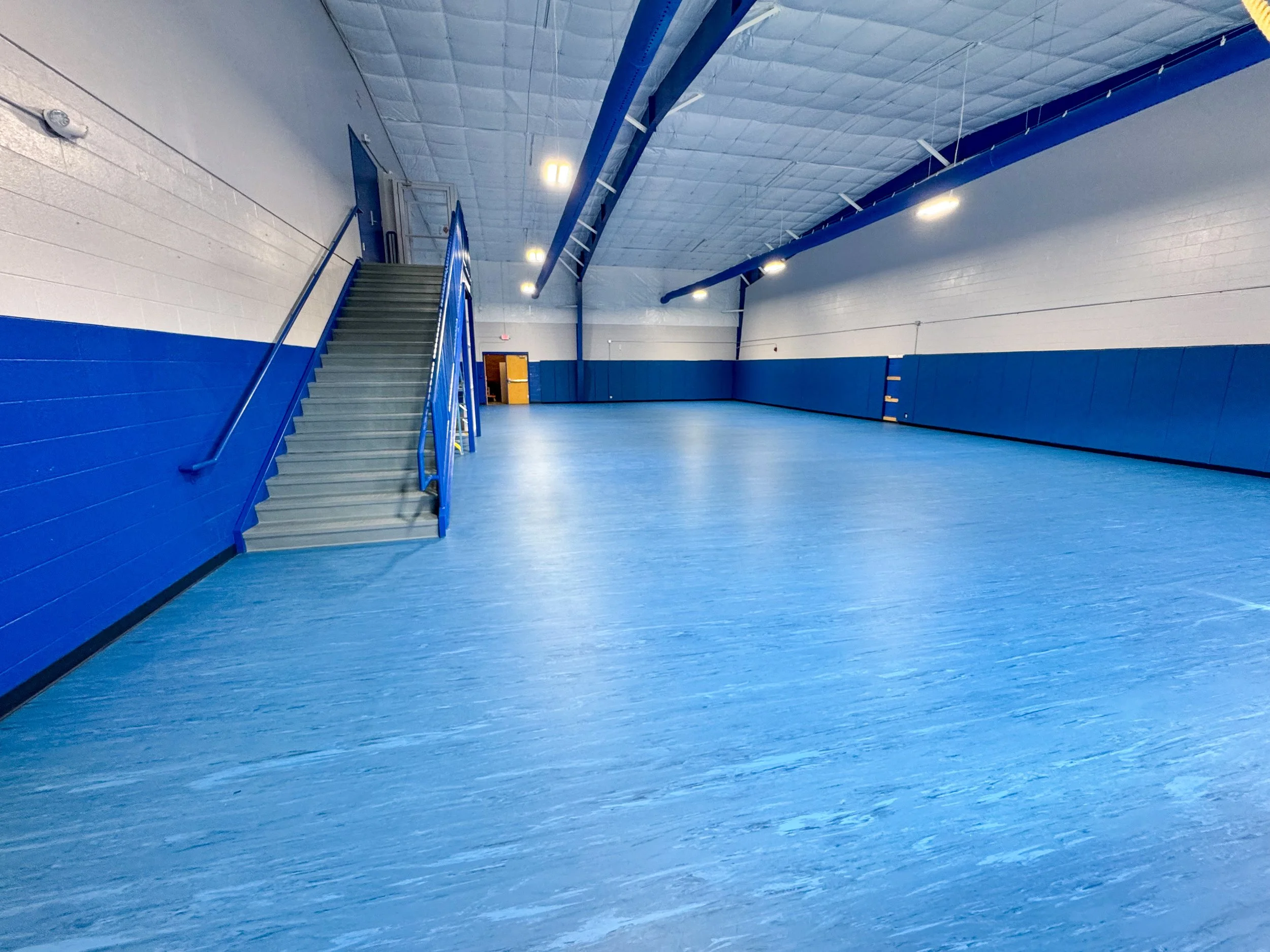 Empty indoor sports gymnasium with blue and white walls, blue floor, and a staircase leading to a door.