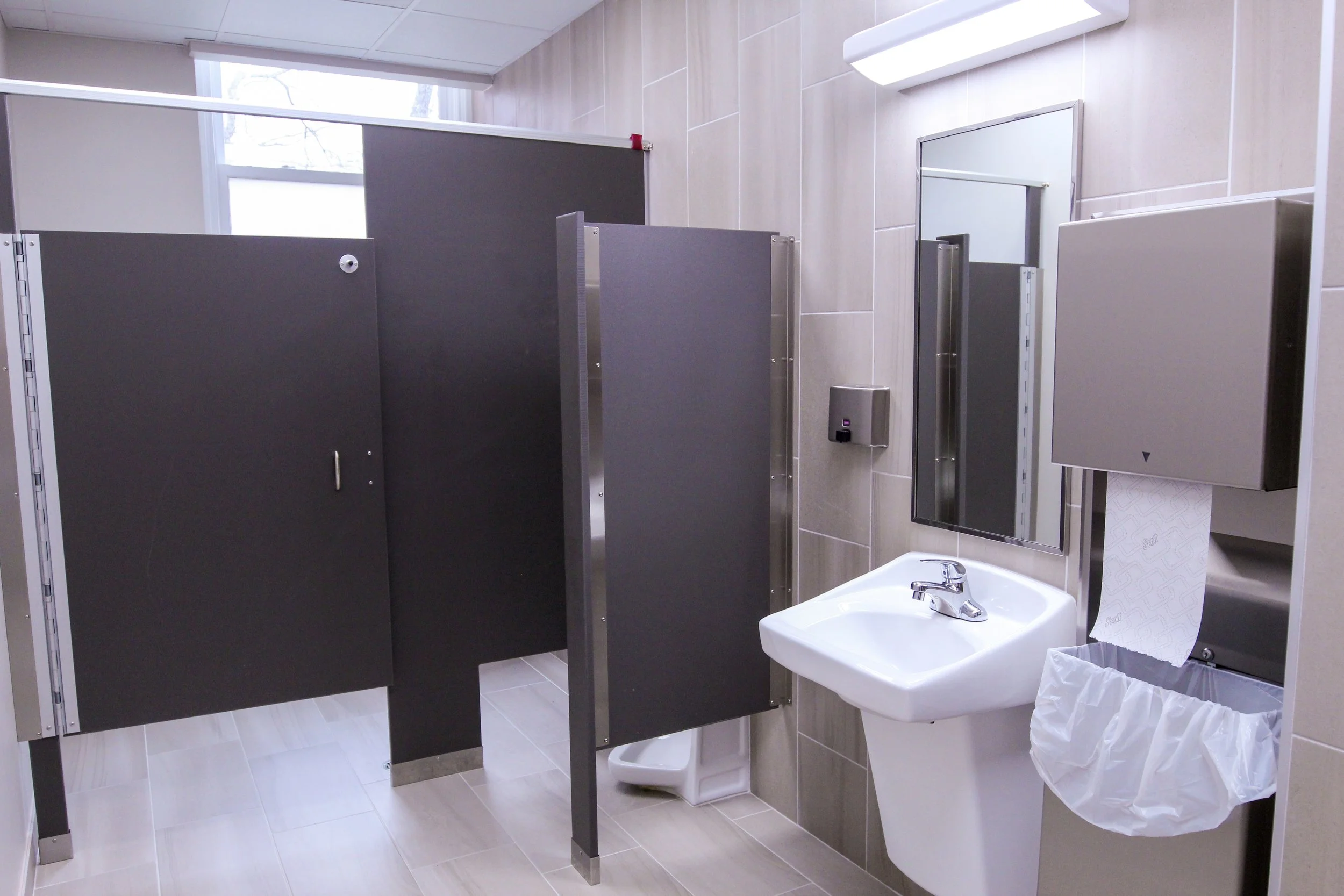 Public restroom with beige tiled walls, a white sink with a faucet, a mirror above, a hand sanitizer dispenser, a paper towel dispenser with a paper towel hanging, and black stall dividers.