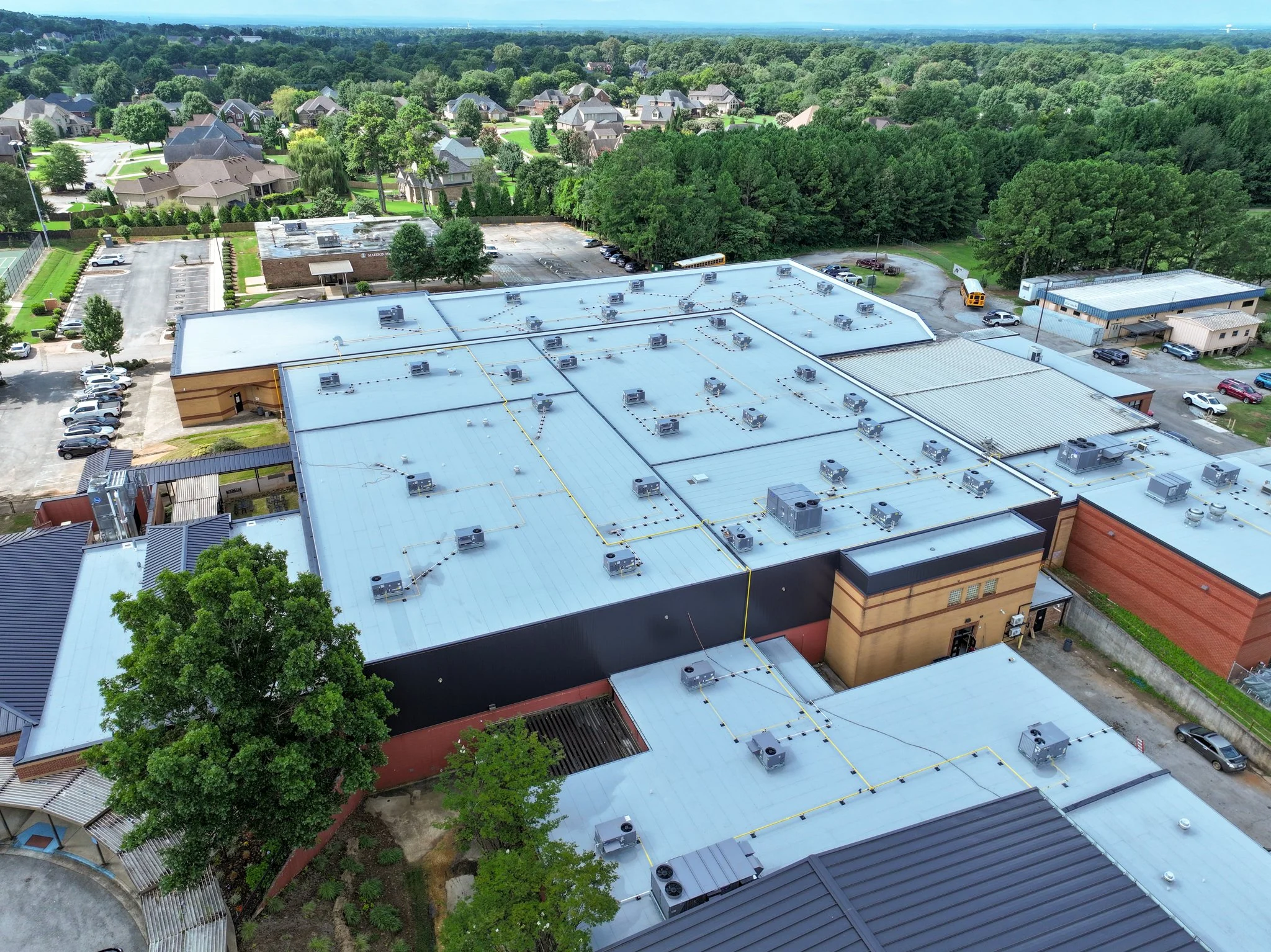Aerial view of Discovery Middle School with a light blue metal roof, numerous HVAC units, a parking lot with cars, and a surrounding green residential neighborhood with trees and houses. Built by P&C Construction. 