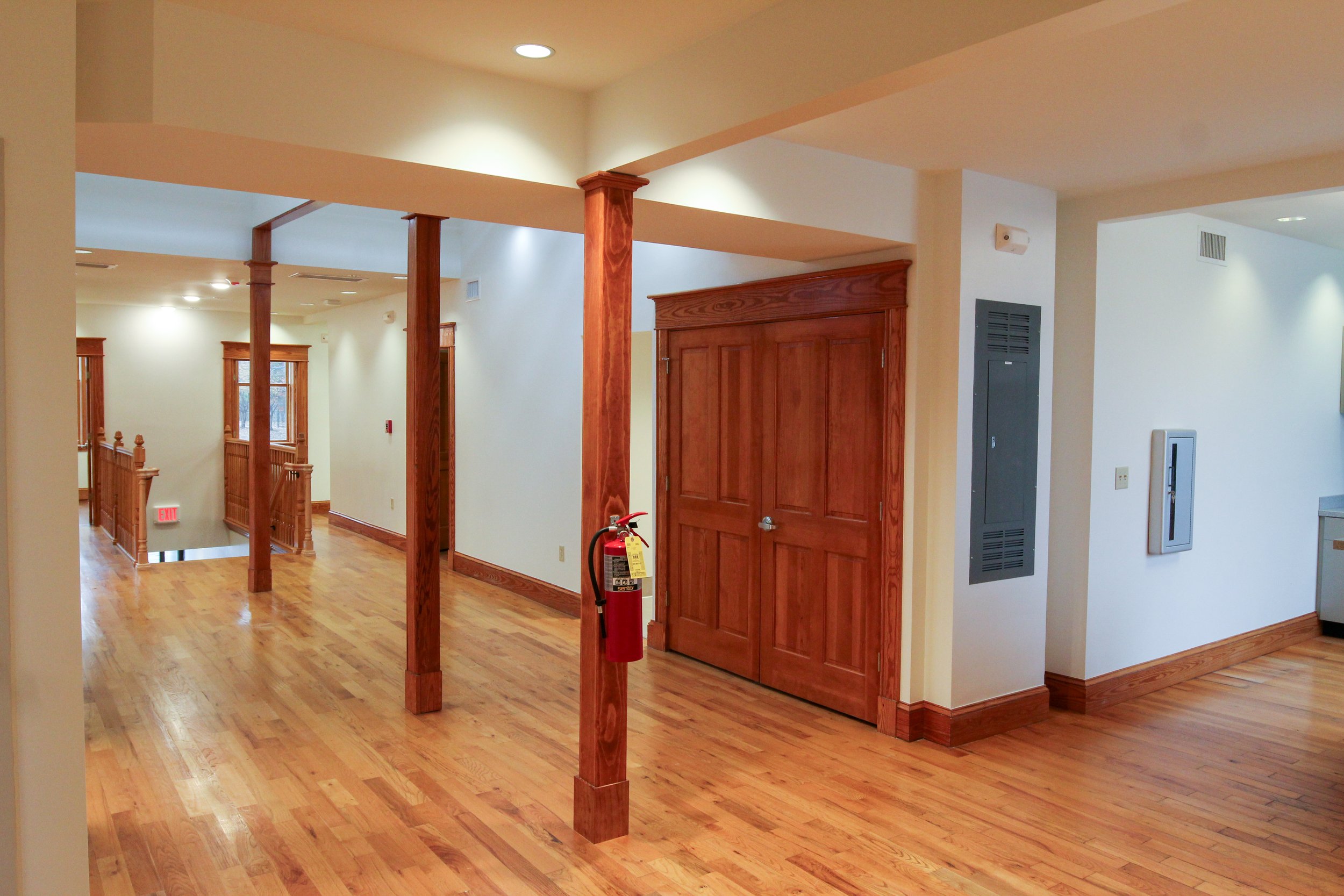 Interior hallway with wooden floors, wooden columns, and doors; fire extinguisher on one column, electrical panels on wall.