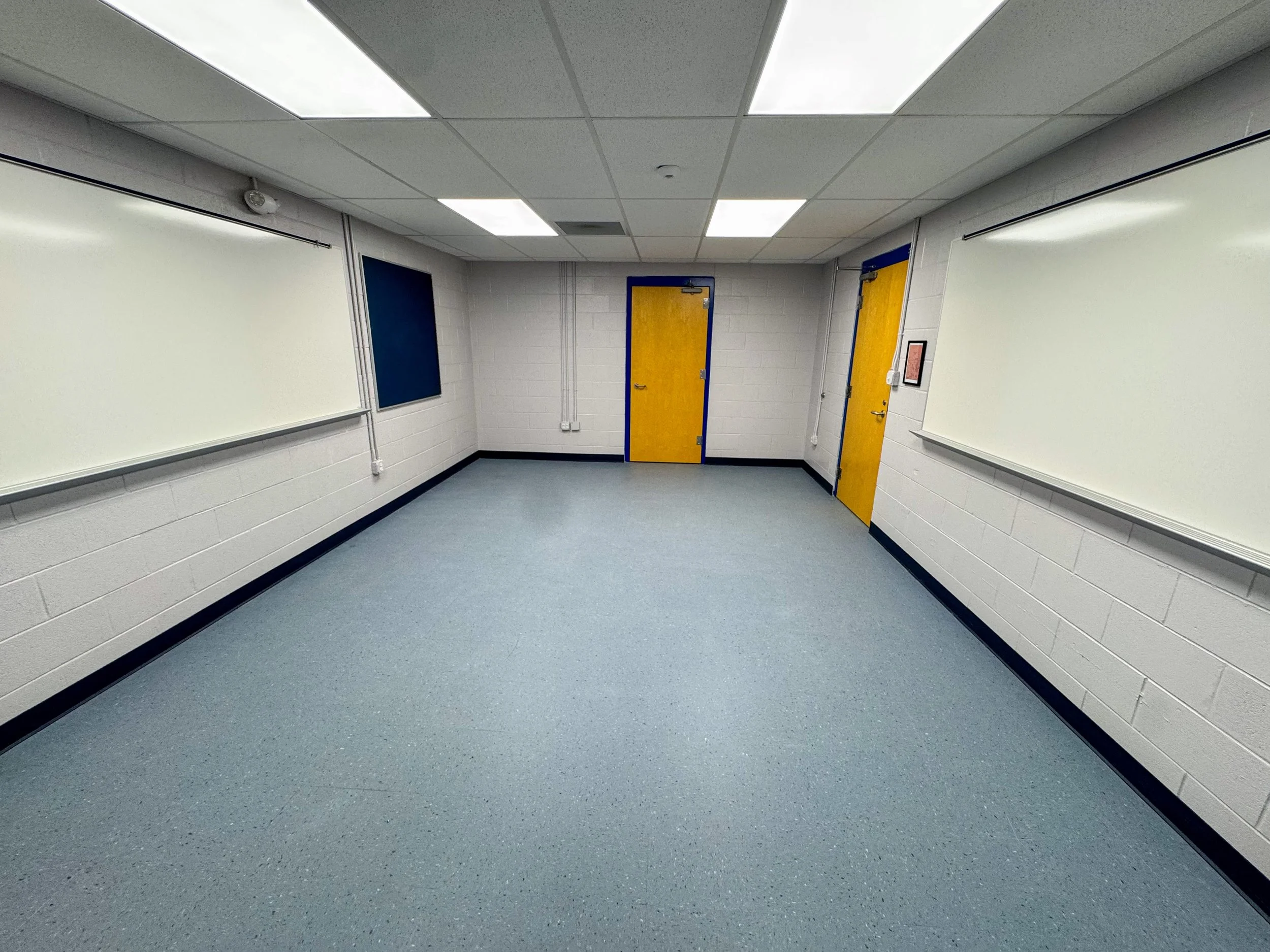 Empty classroom with white brick walls, gray carpeted floor, suspended ceiling with fluorescent lights, two yellow doors with blue frames, two whiteboards on the walls, and a blue bulletin board.