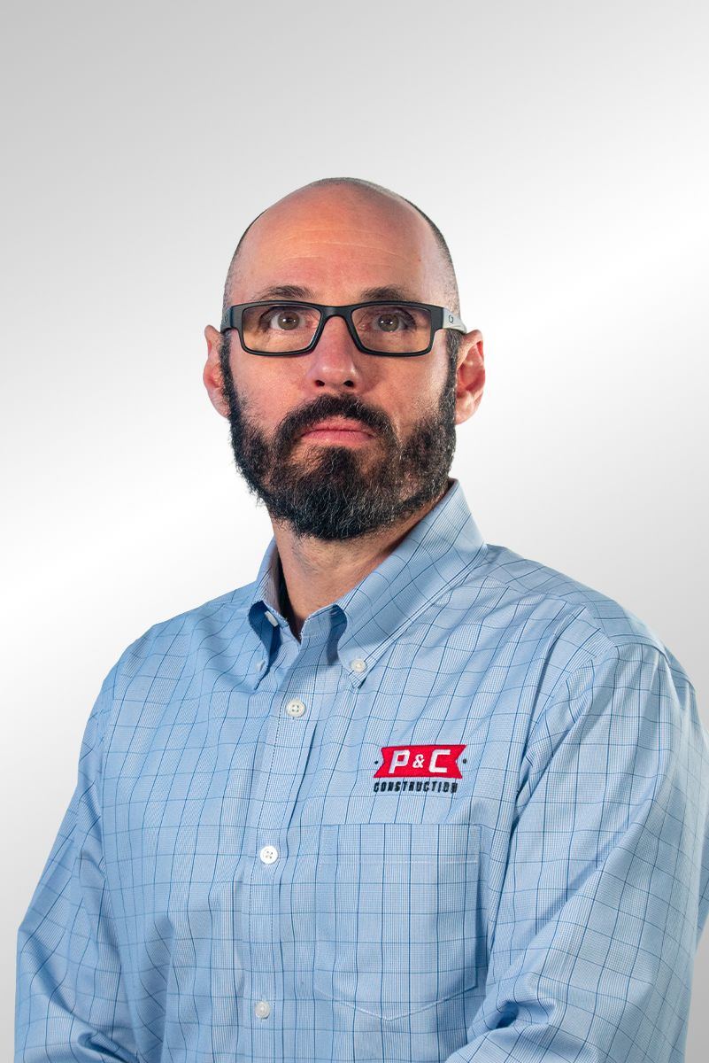 Portrait of a man with glasses, a beard, and a mustache, wearing a white button-up shirt with a subtle checkered pattern, standing against a plain gray background.