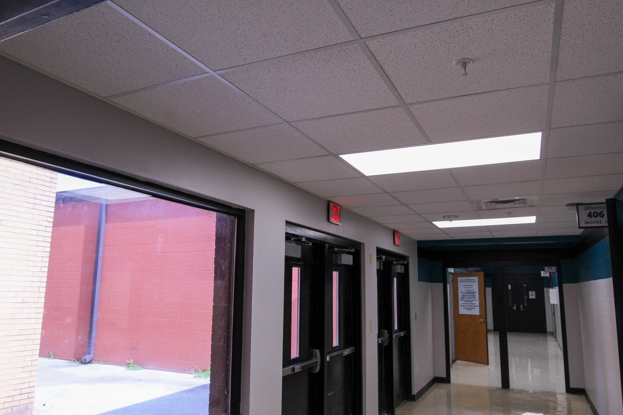 Interior of a building with a drop ceiling, fluorescent lights, large windows, and doors leading outside. An exit sign is visible above the door.