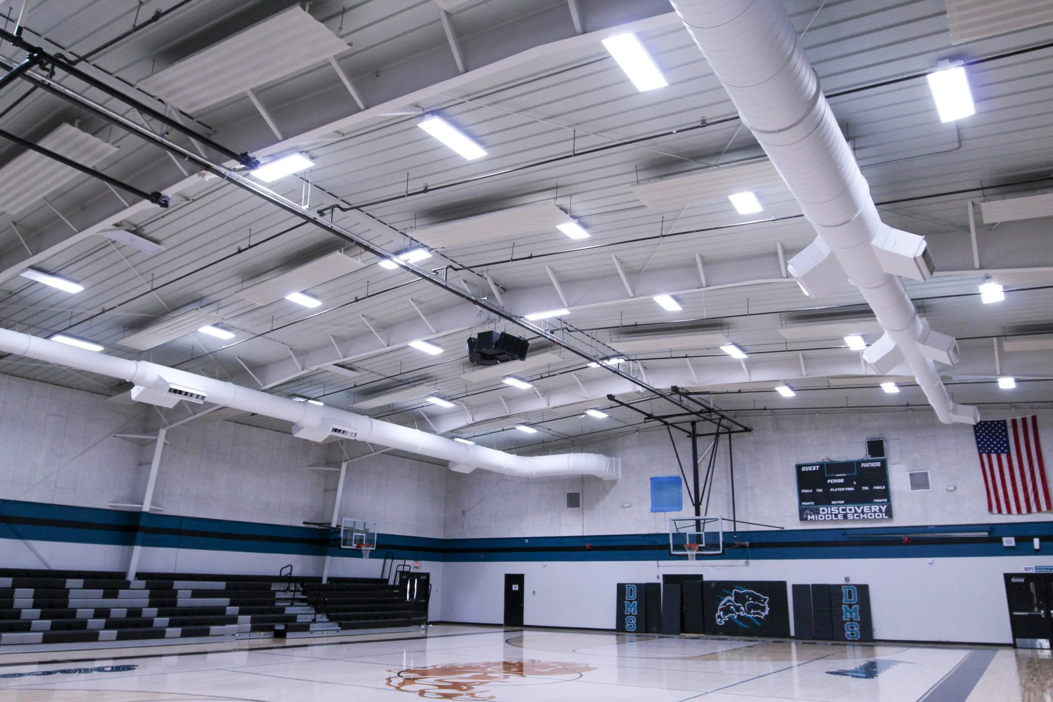 Indoor basketball court with a wooden floor, basketball hoops, and a scoreboard. The gym has high ceilings with white panels, large air ducts, and bright lighting. An American flag is visible on the wall.