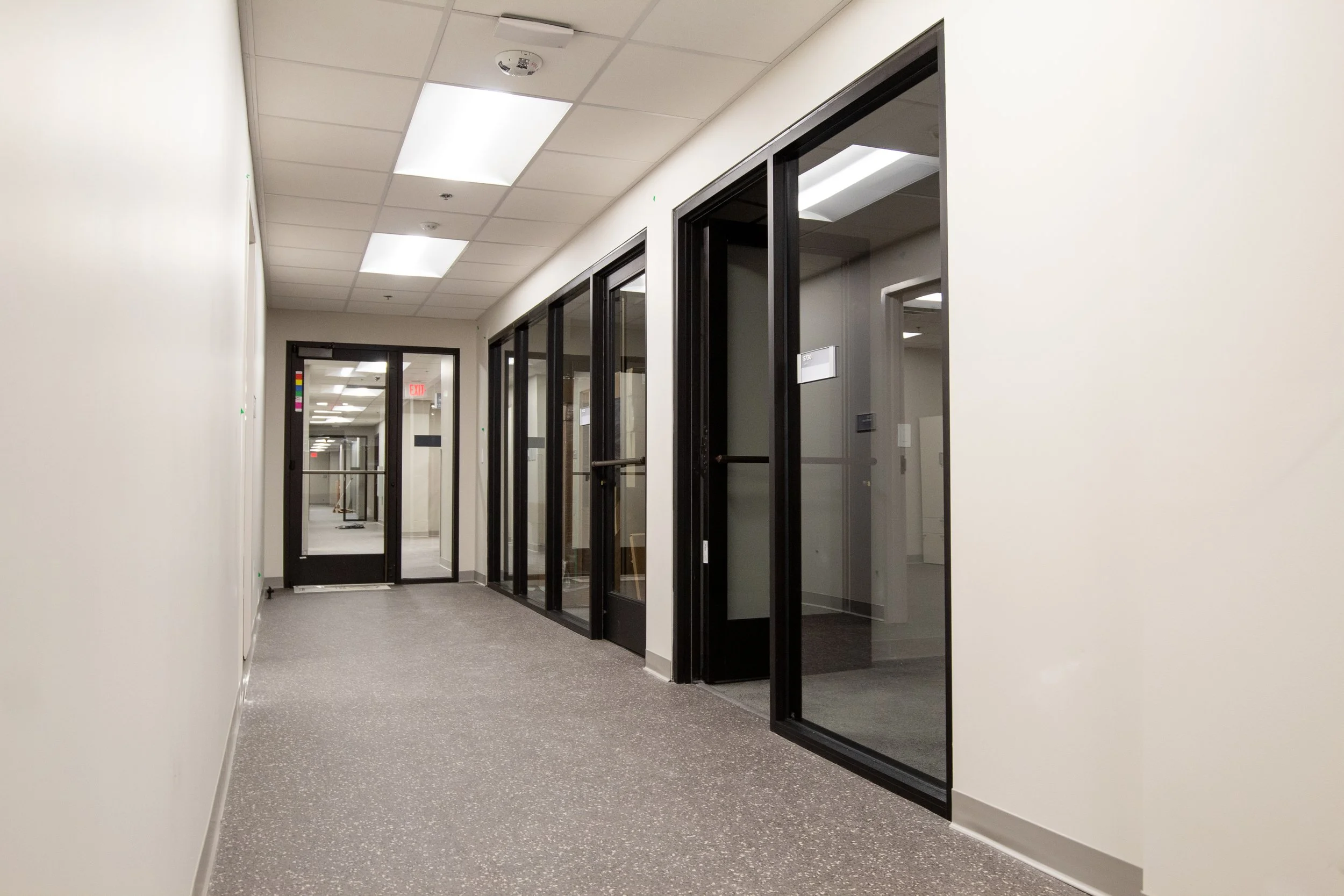 Empty office corridor with glass-walled conference rooms on the right and a closed glass door at the end with an exit sign above it.