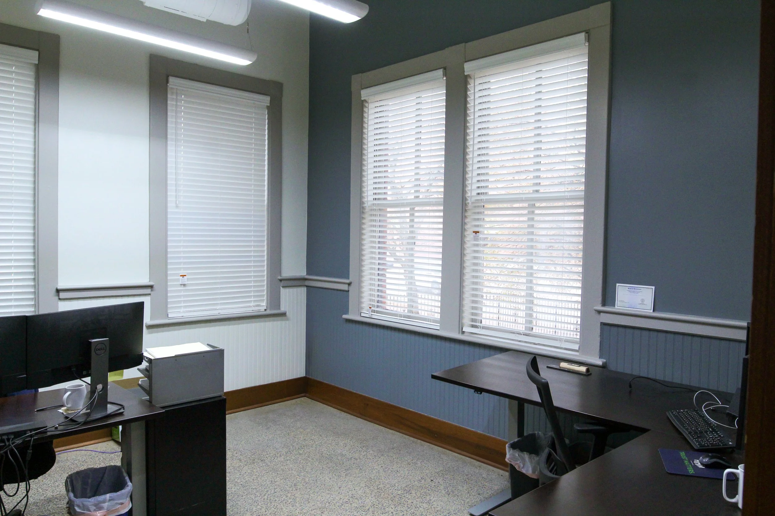 An office with three large windows with white blinds, two desks with computers, chairs, a trash can, and a certificate on the wall.