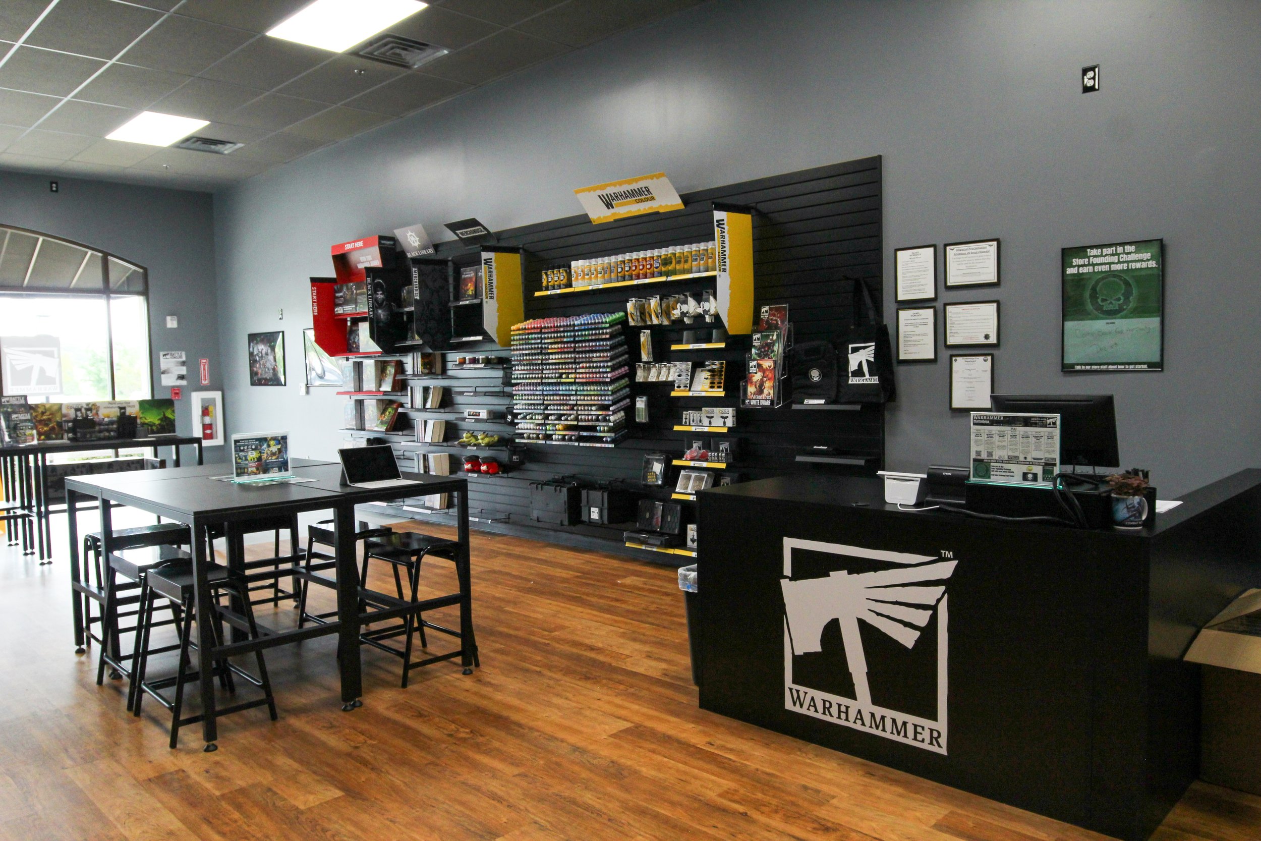 Interior of a store or gaming shop with Warhammer merchandise on black display shelves, tables with chairs, and framed informational posters on a gray wall. Remodeled by P&C Construction, Inc. - Chattanooga, Tn.