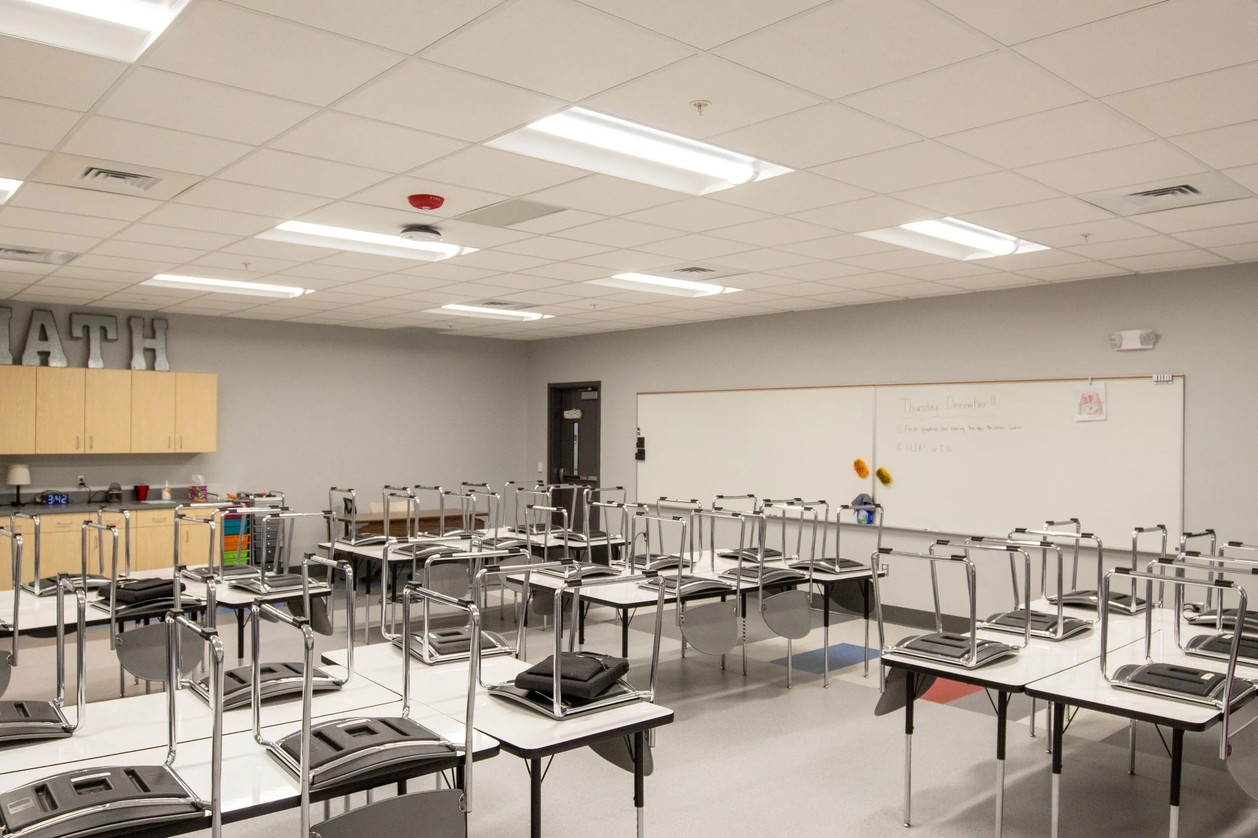 Empty classroom with chairs stacked on desks, whiteboard, cabinets, and ceiling lights.