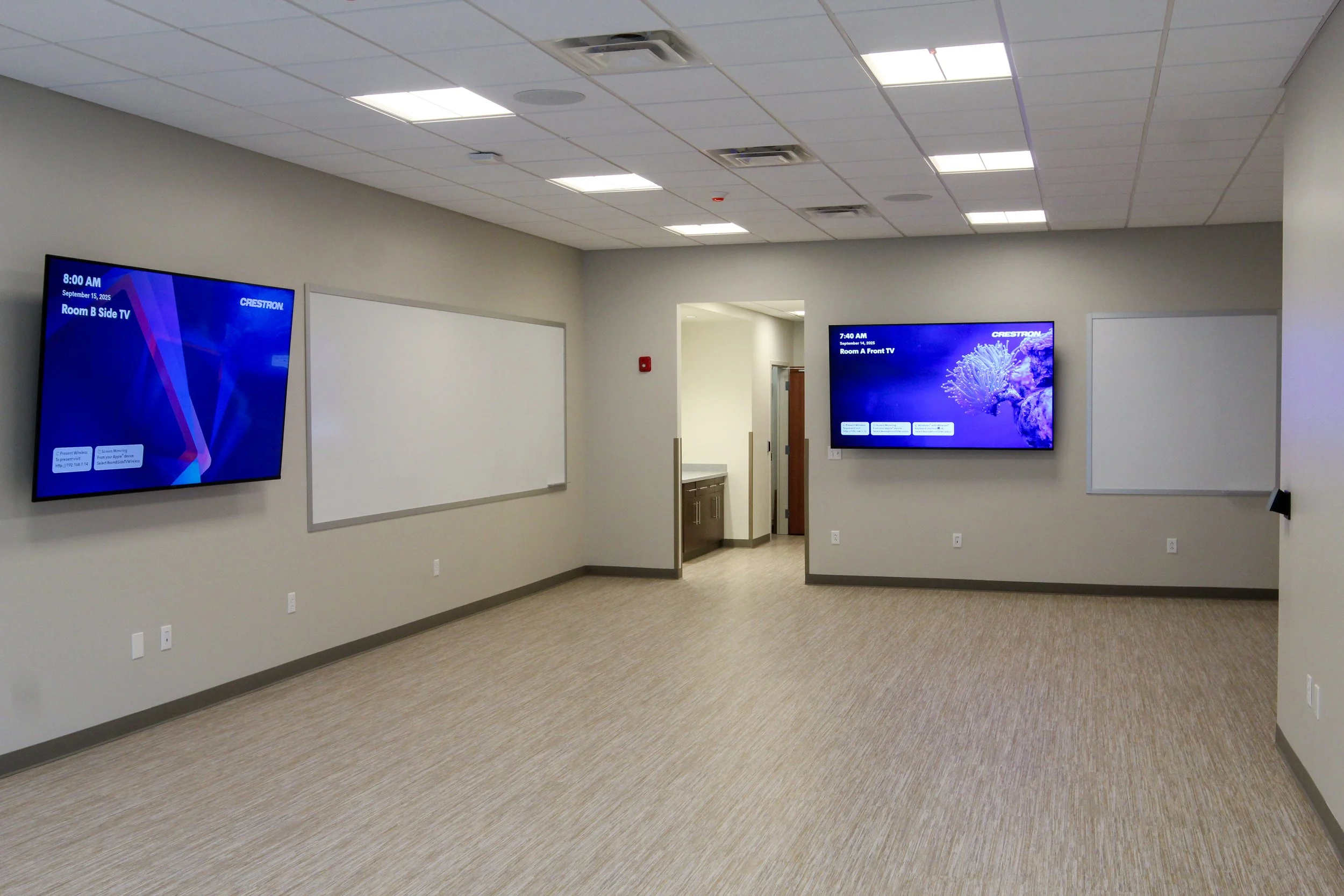 Empty conference or meeting room with two wall-mounted screens showing digital displays, whiteboards, and beige carpeted flooring.