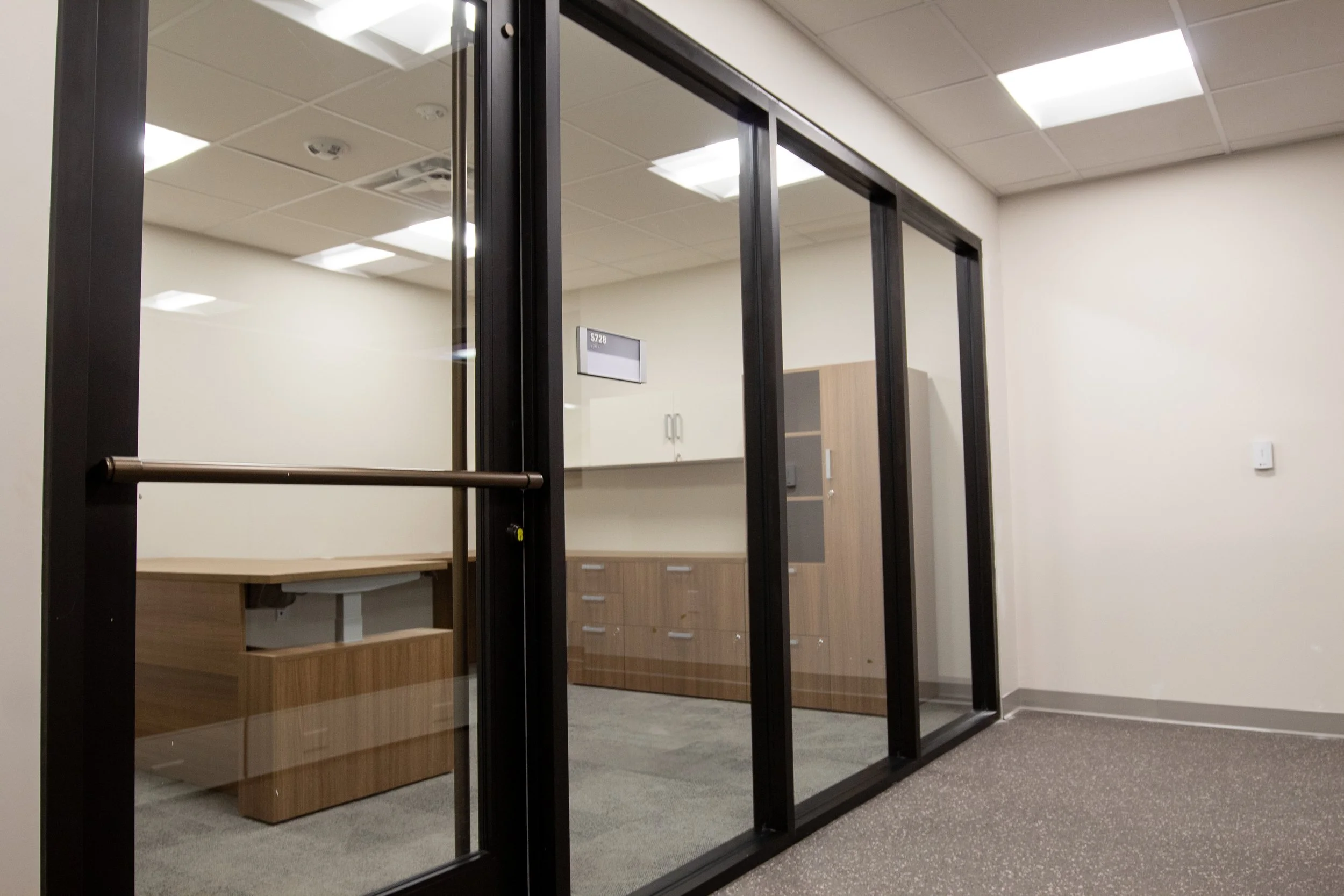 Empty office conference room with glass walls, a wooden desk, cabinet, and ceiling lights.