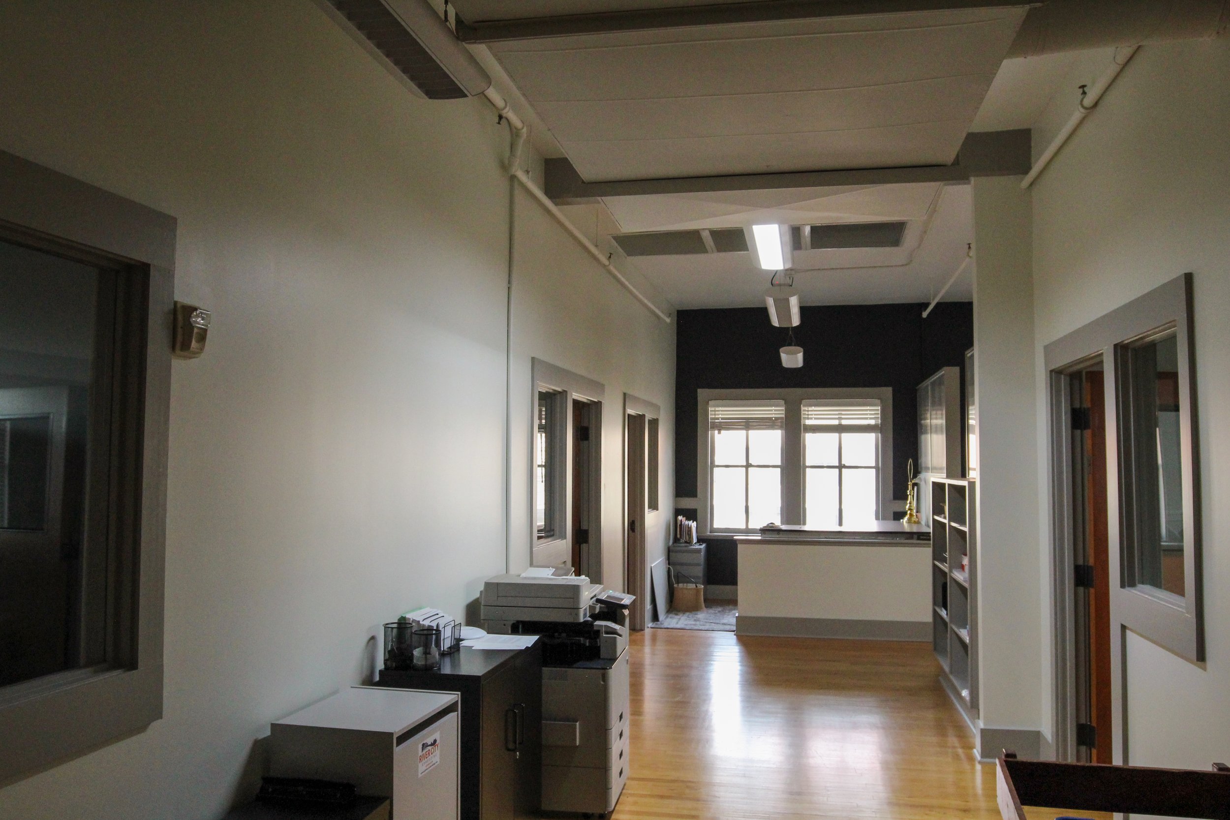 Indoor office space with light-colored walls, hardwood floors, desks, and windows at the end of the room.